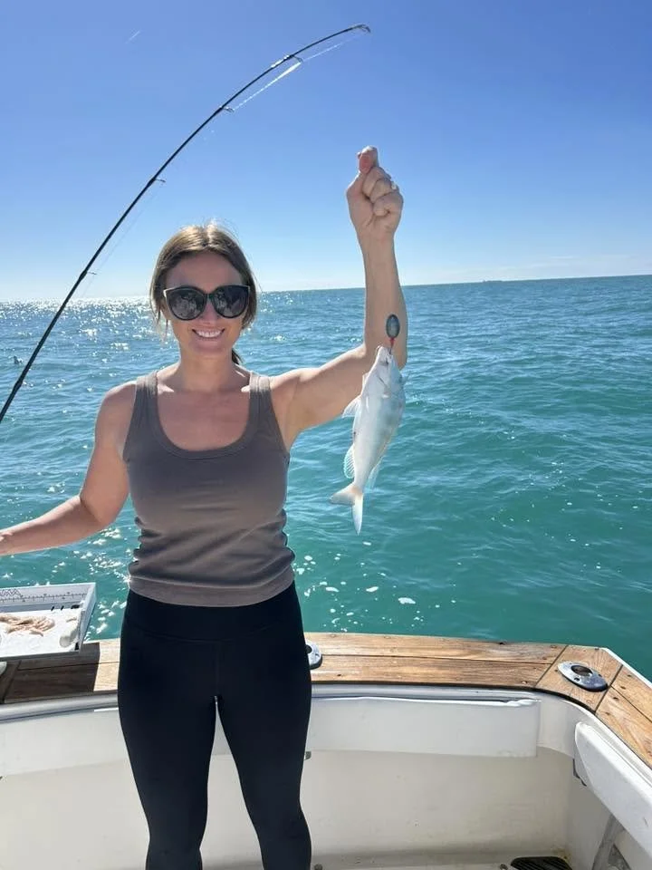 Smiling woman wearing sunglasses proudly holding up a freshly caught fish aboard a boat on the Gulf of Mexico.