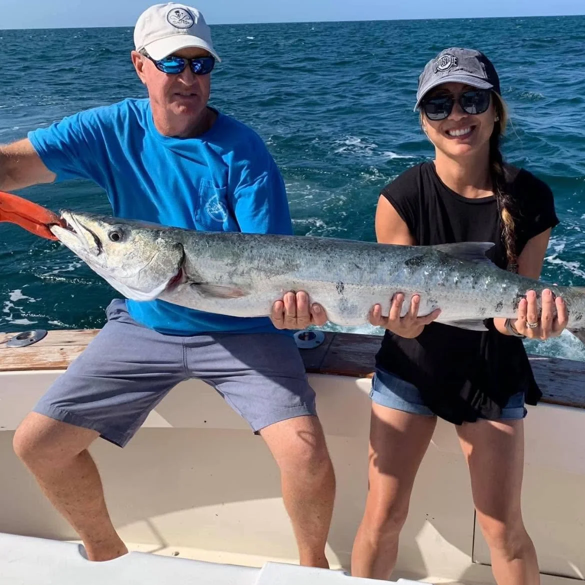 Two smiling anglers holding a large silver fish horizontally aboard a fishing charter boat.