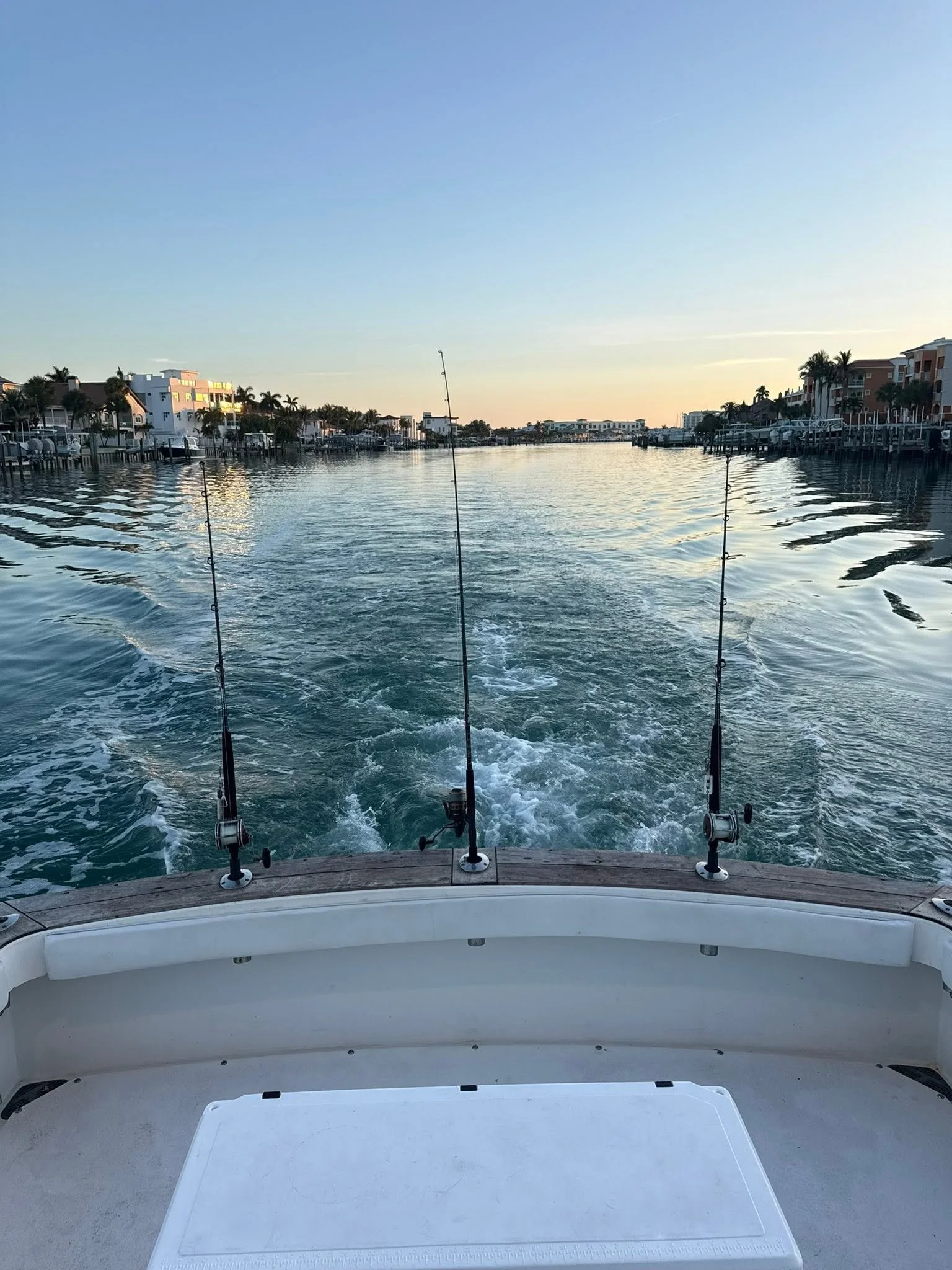 View from the stern of a fishing boat leaving a wake along a calm coastal waterway lined with waterfront homes and docks.