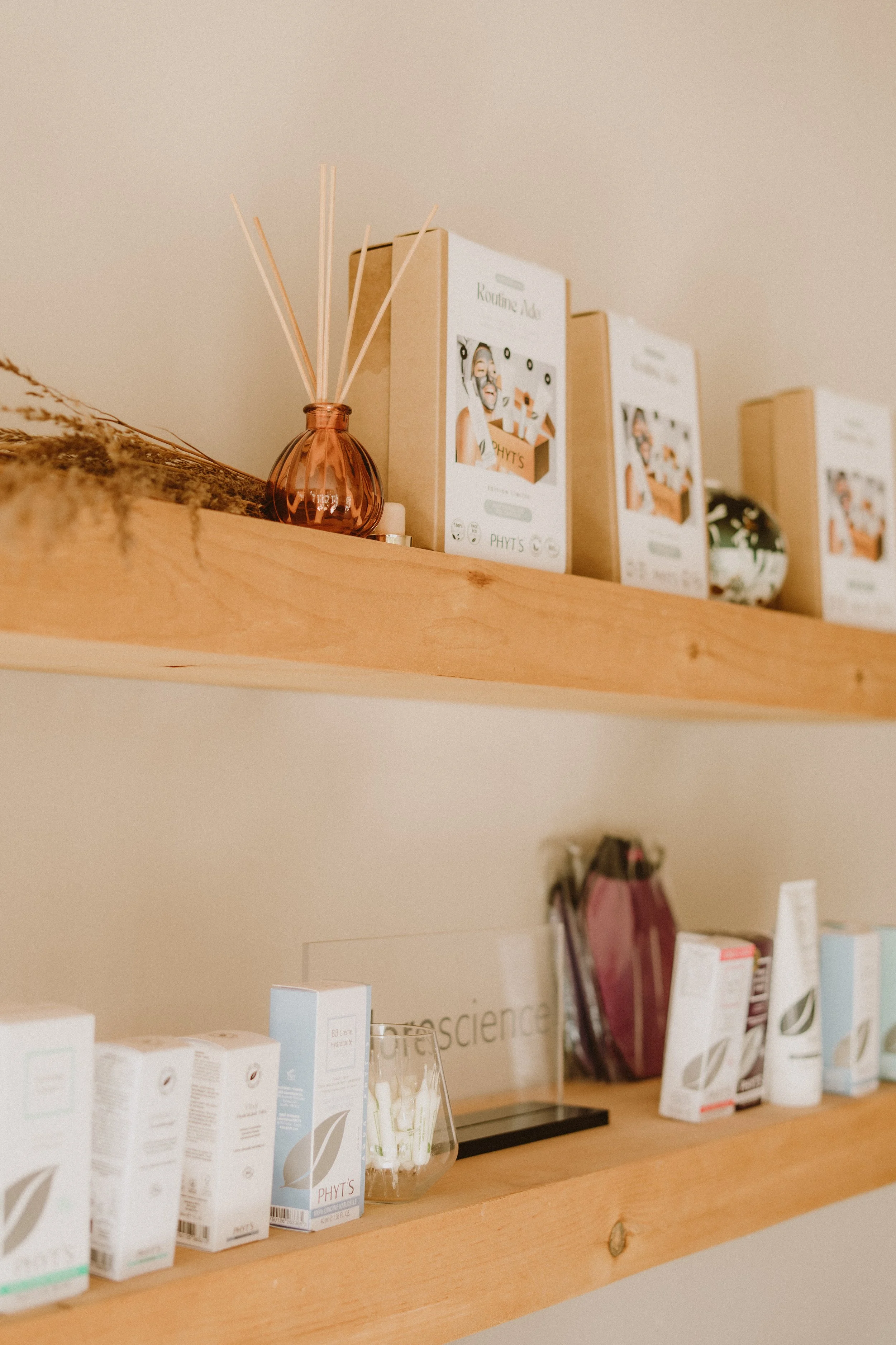 Près d'une étagère en bois, plusieurs produits de soins de la peau et de bien-être de la marque Phyt's, notamment des boîtes, des tubes, et une petite jarre avec des bâtonnets parfumés.