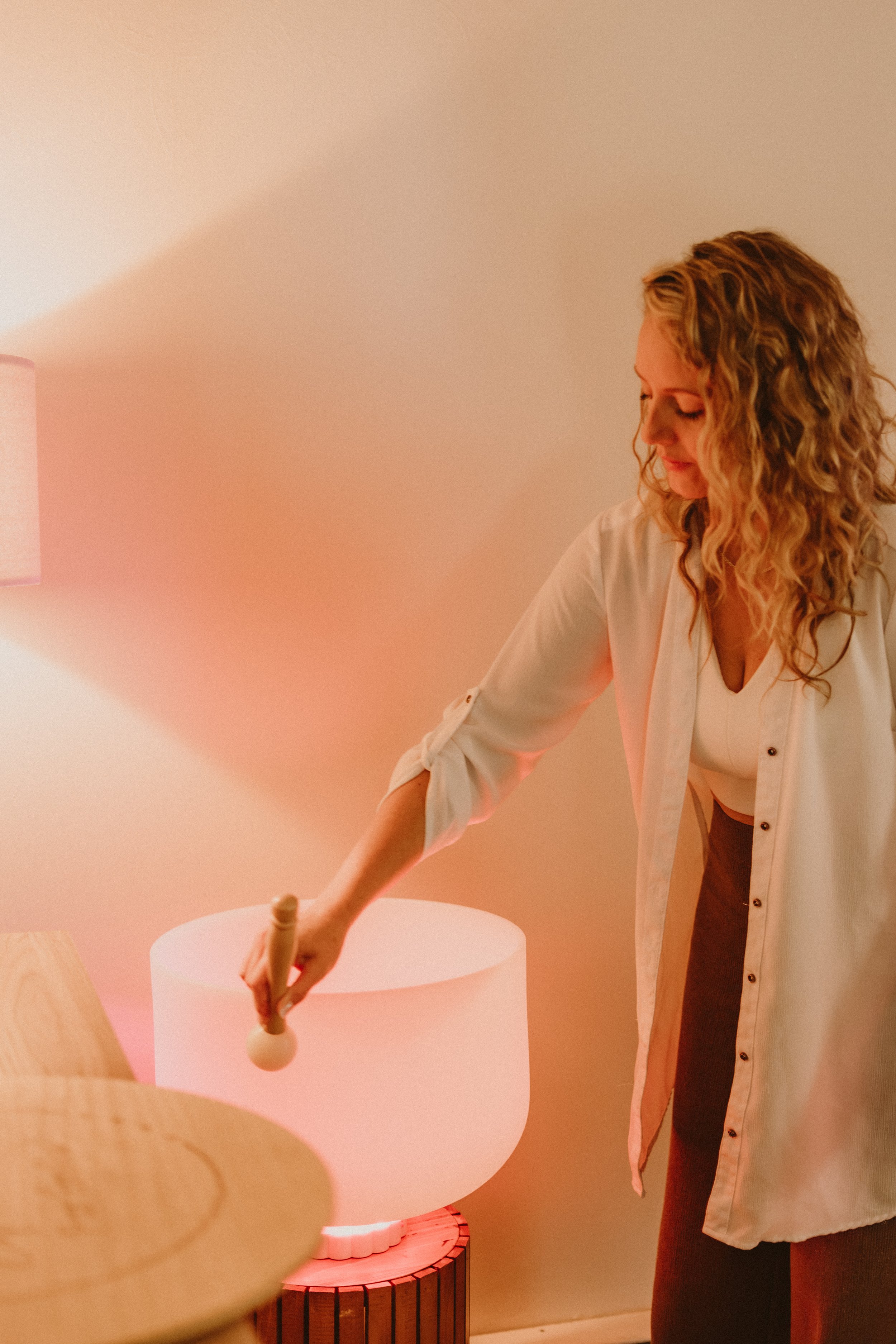 Une femme aux cheveux bouclés et vêtue d'une chemise blanche, qui semble jouer ou manipuler un objet en bois dans une pièce aux murs rosés, avec un lampadaire à l'arrière.