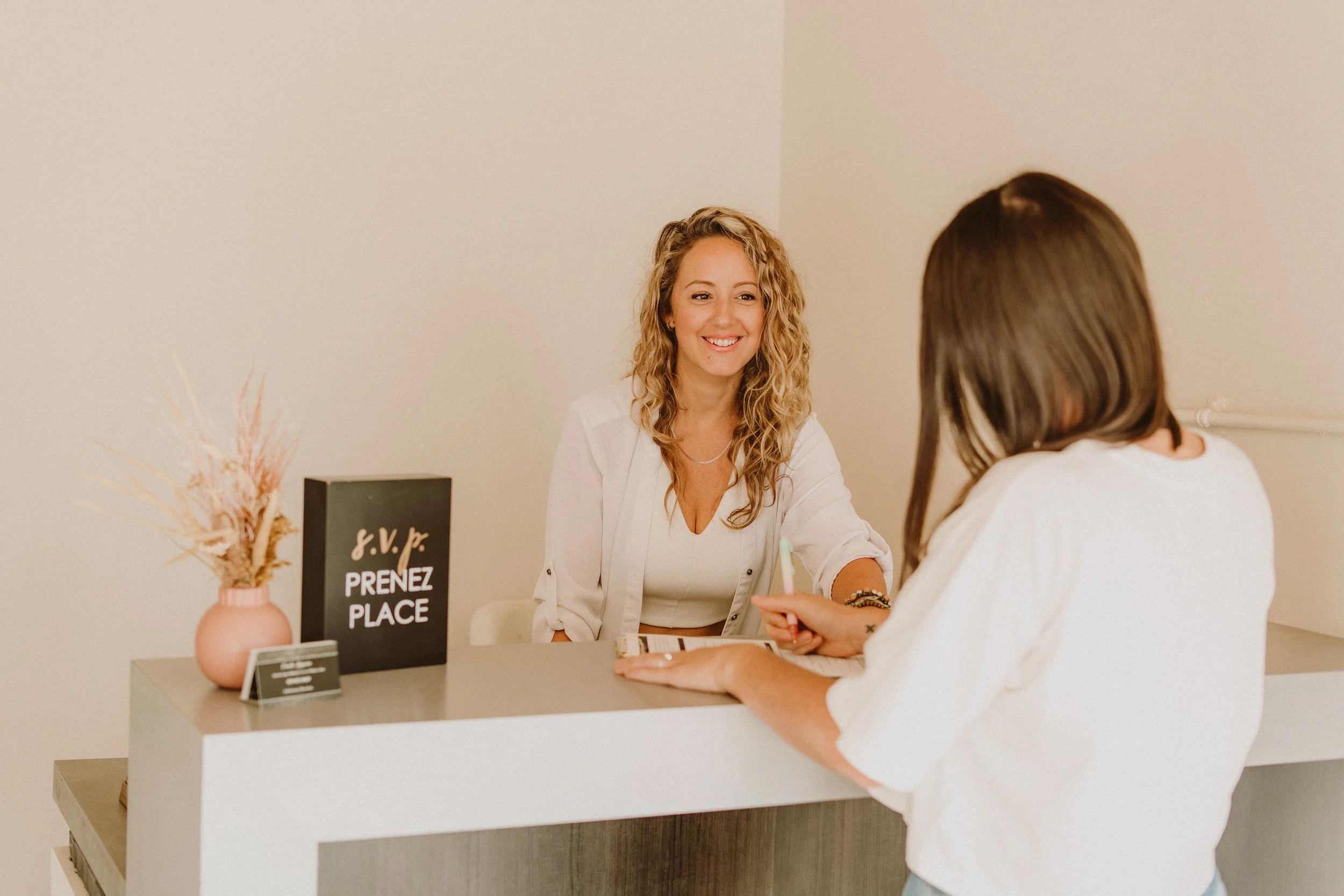 Émilie Giguère discute avec une personne assise à un comptoir chez Espace M, clinique esthétique de Lachute. Sur le comptoir, un vase avec des fleurs séchées et un panneau qui invite à prendre place.