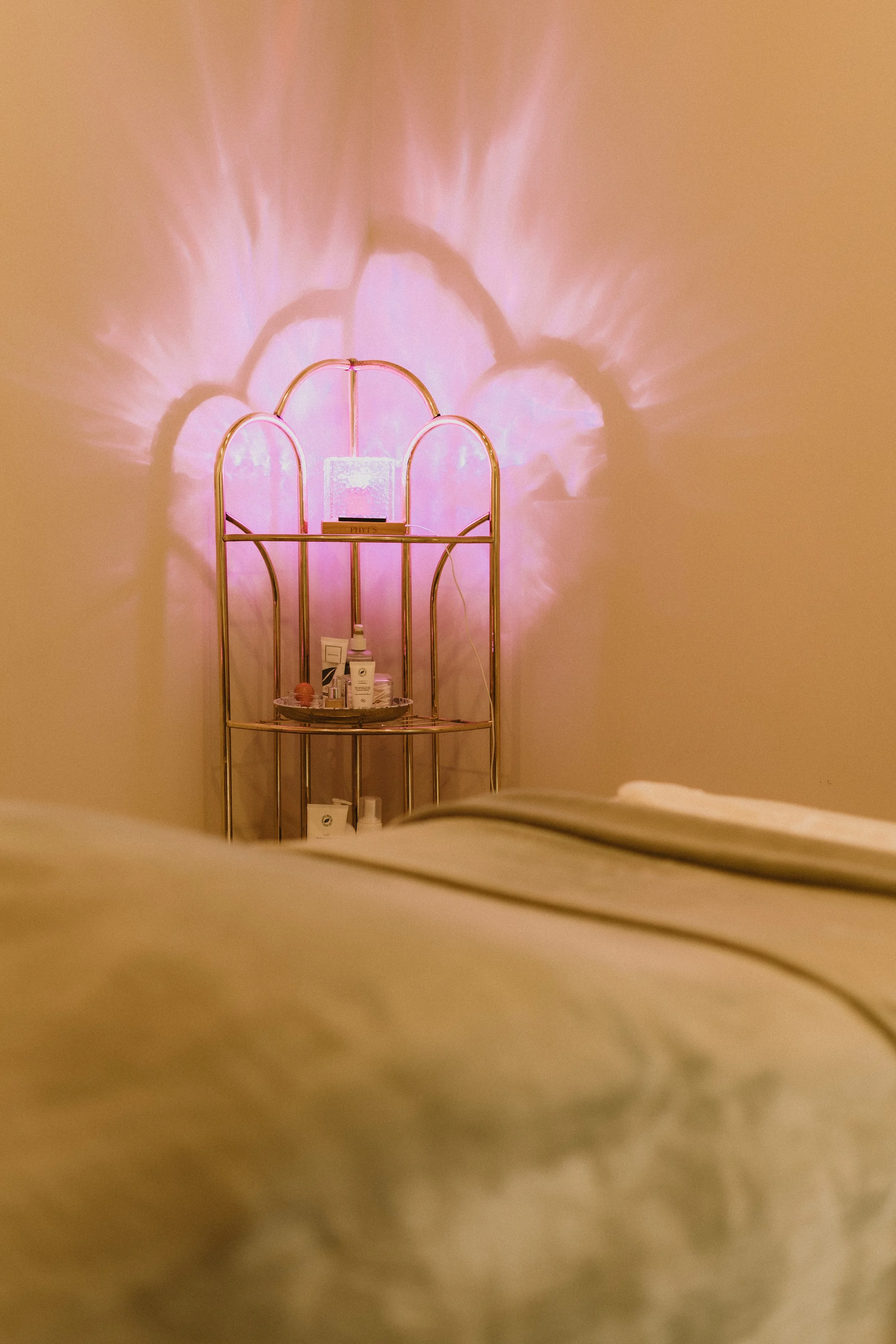 Une étagère dorée avec divers produits de beauté ou soins, éclairée par une lumière rose qui projette des motifs en forme d'arcs sur le mur blanc derrière.
