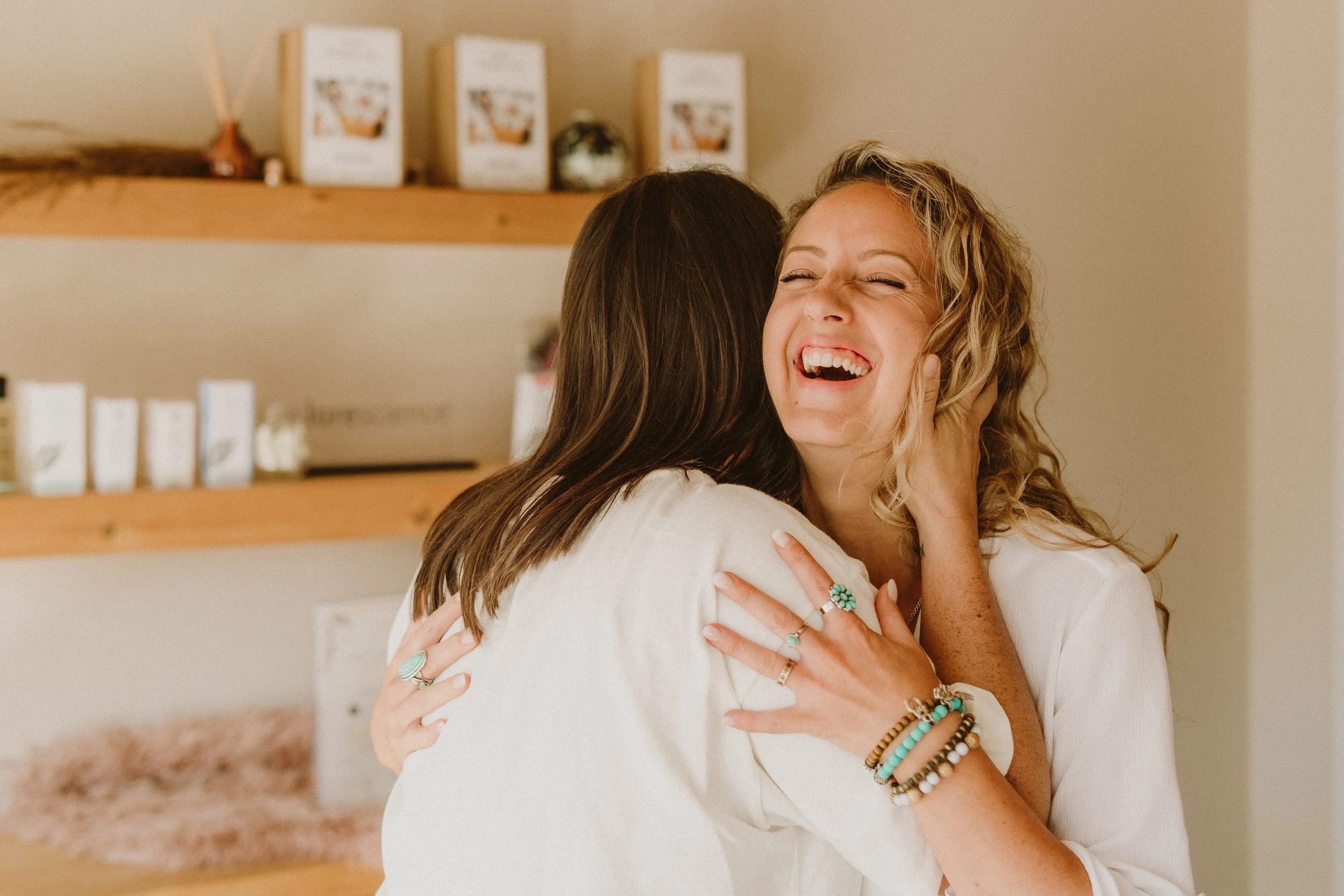 Deux femmes s'embrassent et sourient dans une ambiance chaleureuse, décorée avec des livres et des objets dans un intérieur.
