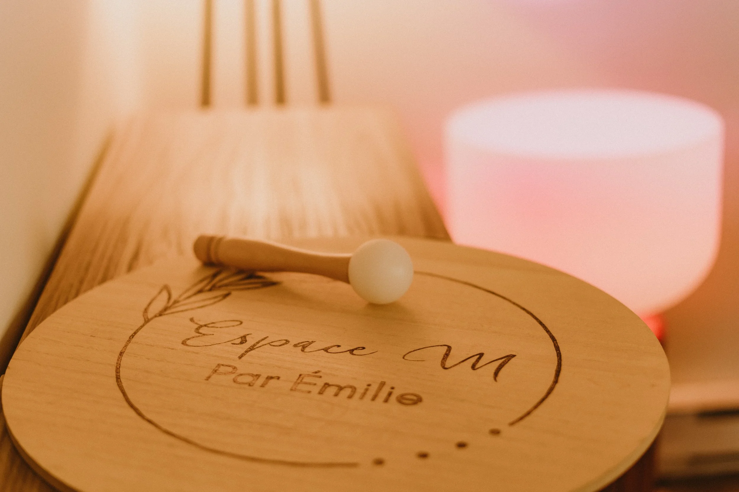Une boîte en bois avec un couvercle gravé portant le message 'Espace Aimé pour Famille', sur laquelle repose un petit objet en bois ressemblant à une manette ou un outil rond. En arrière-plan, une lampe en bois avec un abat-jour en tissu rose donne une lumière chaleureuse à la scène.