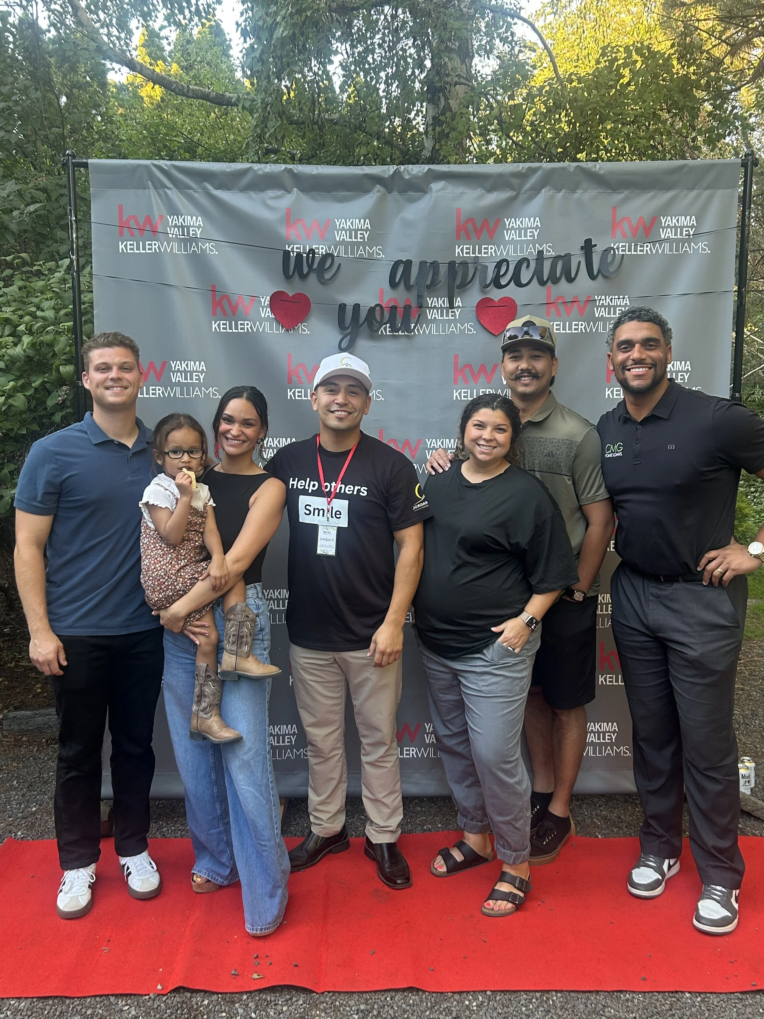 A group of seven people, including a small girl, standing on a red carpet in front of a backdrop with the logos of Keller Williams and Yakima Valley, at an outdoor event. The people are smiling and posing for the photo.