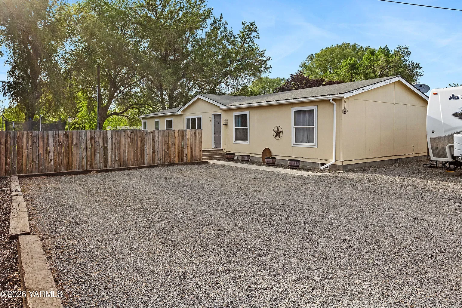 A single-story yellow house with a gravel driveway, a wooden fence, potted plants, and a star wall decoration. Large trees and a blue sky in the background.