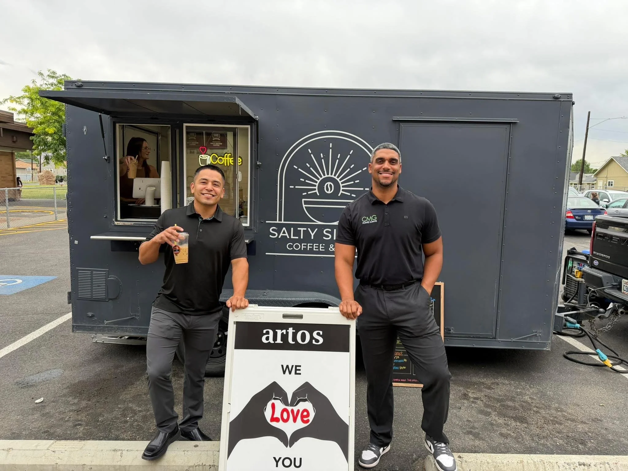 Two men in black shirts standing outside a mobile coffee shop, smiling at the camera. One is holding a coffee drink. A sign in front reads "artos" and smaller text beneath says "We Love You." In the background, a woman inside the coffee shop is pouring a drink.