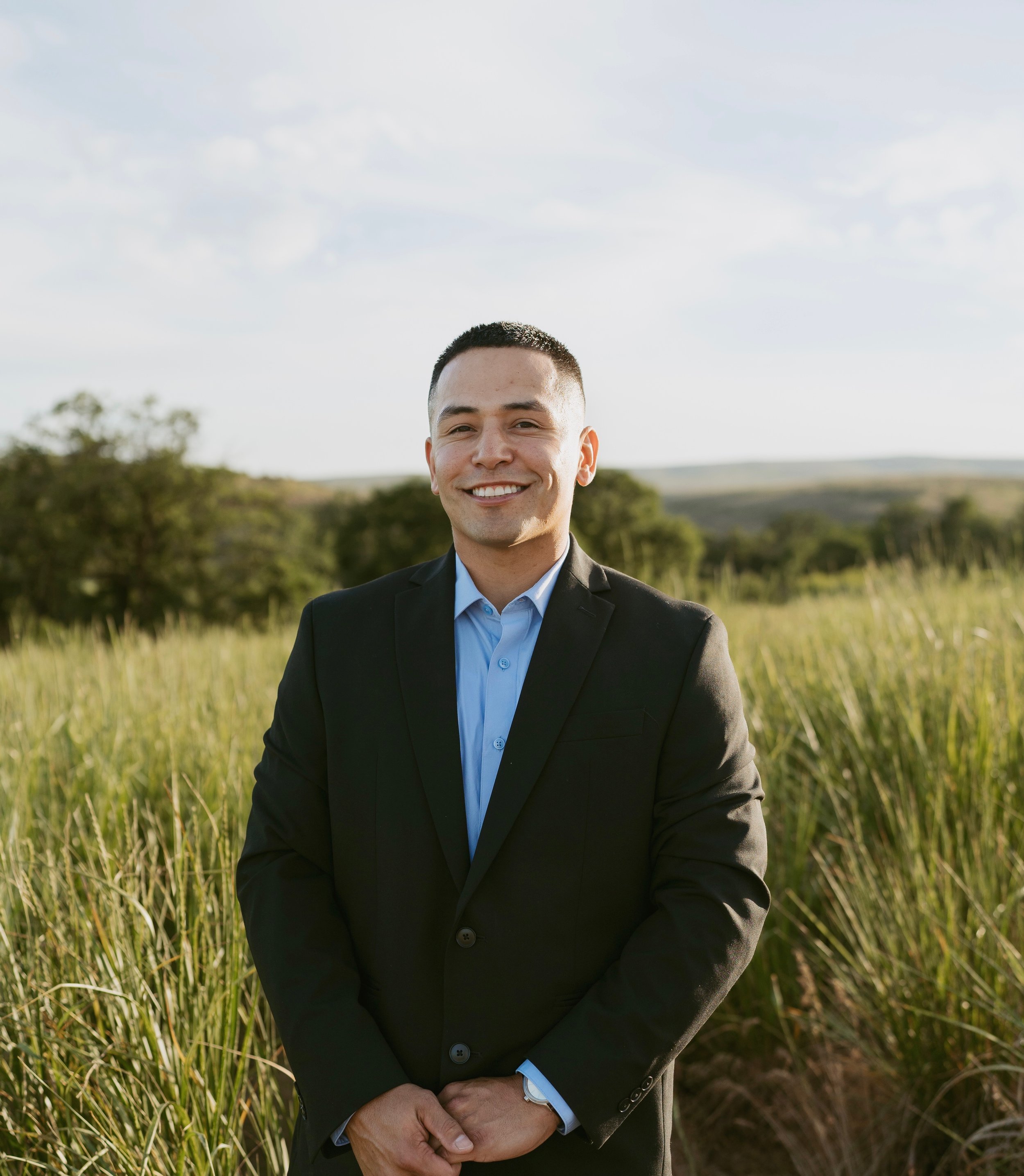 A young man in a black suit with a light blue shirt, smiling, standing outdoors in a field with green grass and trees, under a partly cloudy sky.
