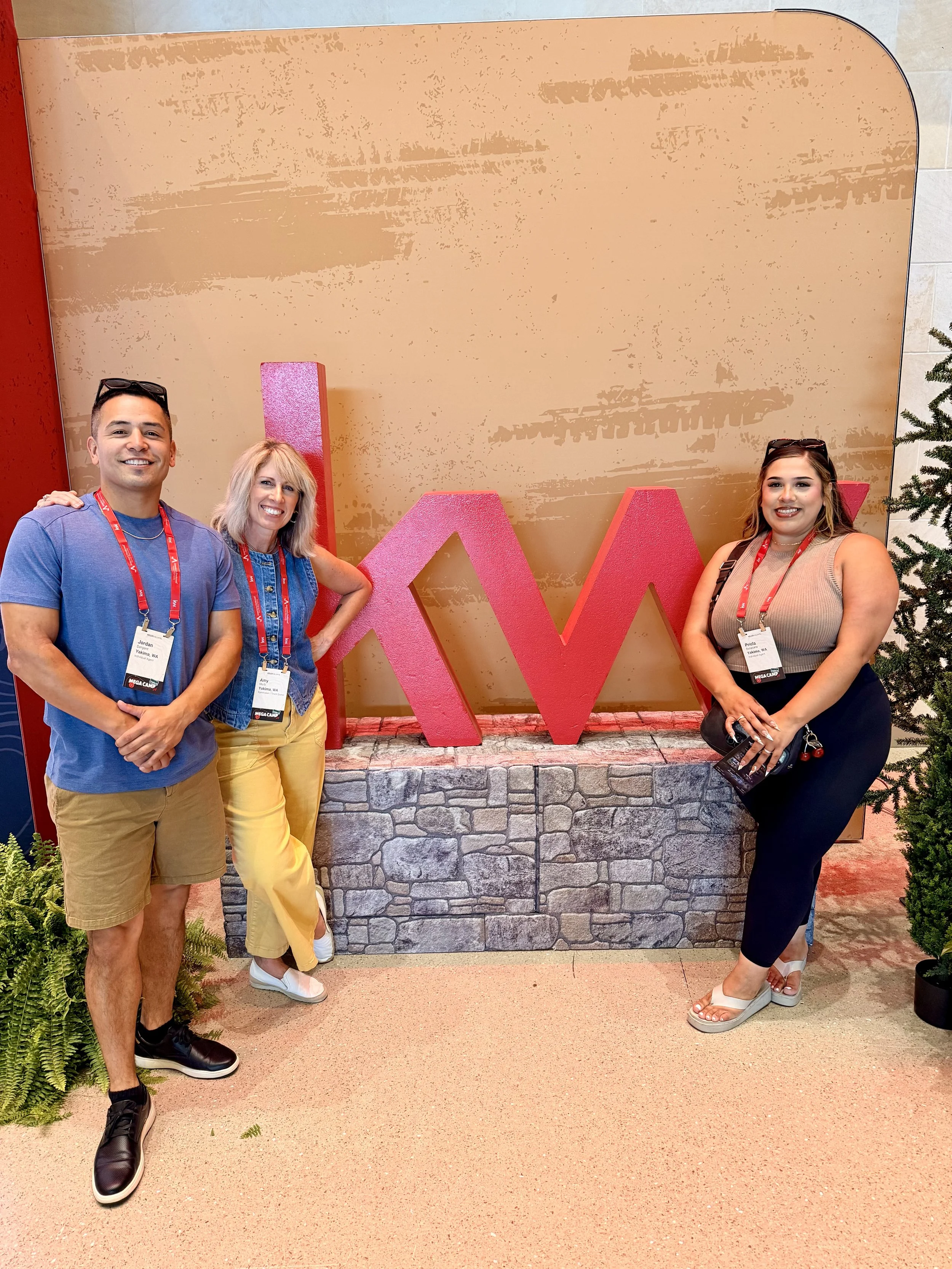 Three people standing in front of a large pink letter 'W' and a yellow wall, smiling and posing for the photo. The person on the left is a man wearing a blue t-shirt, khaki shorts, and black sneakers. The person in the middle is a woman with blonde hair, wearing a denim vest, yellow pants, and white sneakers. The person on the right is a woman with brown hair, wearing a beige sleeveless top, black pants, and white sandals.
