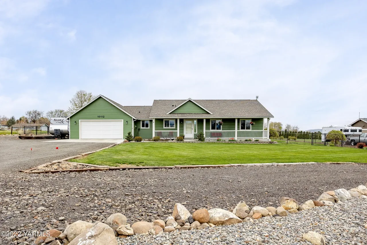 Green house with a front porch and white garage door, surrounded by a lawn and rocks, under a cloudy sky.