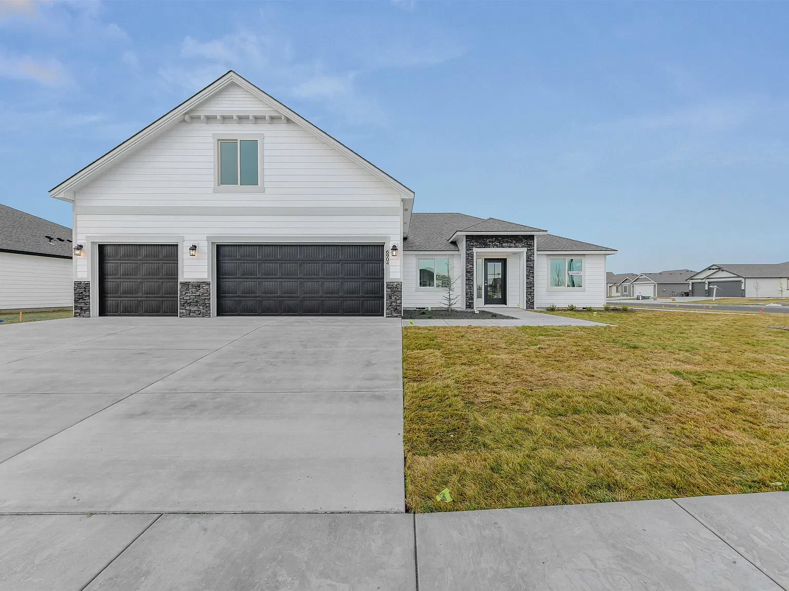 Modern two-story house with white siding, black garage doors, and a small front porch, situated on a lawn with a concrete driveway and a clear sky in the background.
