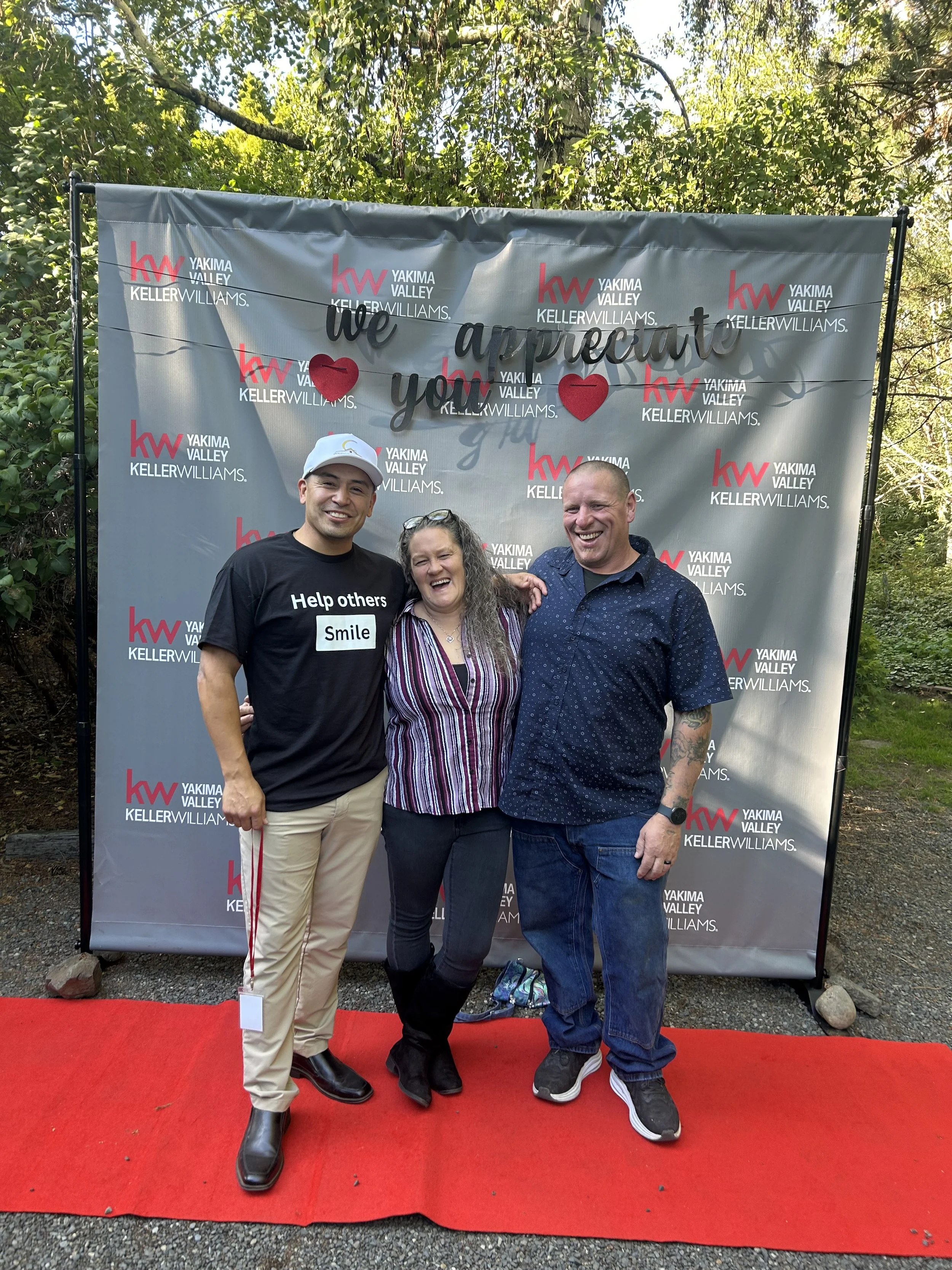 Three people standing on a red carpet in front of a backdrop with the logos of Keller Williams Realty and Yakima Valley. They are smiling and celebrating at an outdoor event during daylight, surrounded by trees.