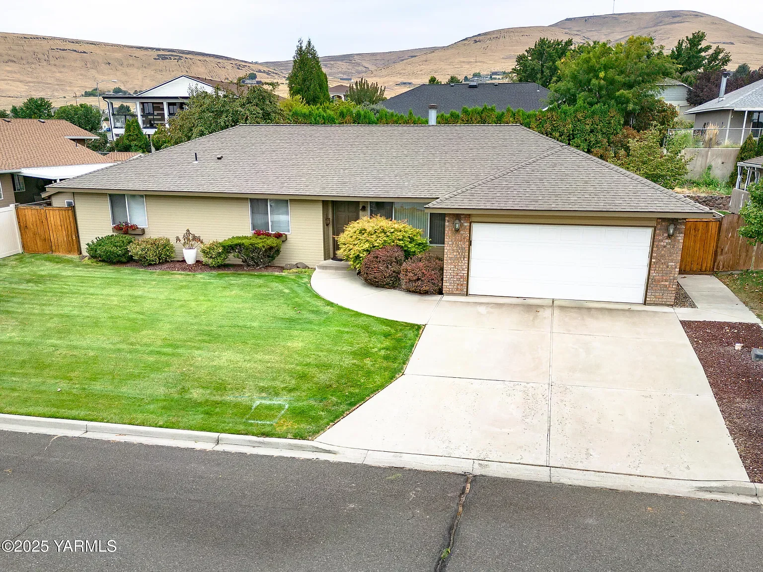 Single-story house with beige siding and brick accents, a white garage door, a well-maintained lawn, and a concrete driveway, with hills and neighboring houses in the background.
