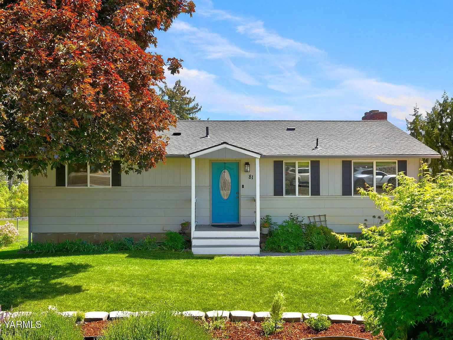 A single-story house with white siding, black shutters, and a gray roof. The front door is bright blue with an oval glass window. A small porch with white stairs leads up to the door. There are two large windows on either side of the house, with a tree on the left and a bush on the right. The house is surrounded by a green lawn and a garden bed with flowers in the front. The sky is blue with some clouds.