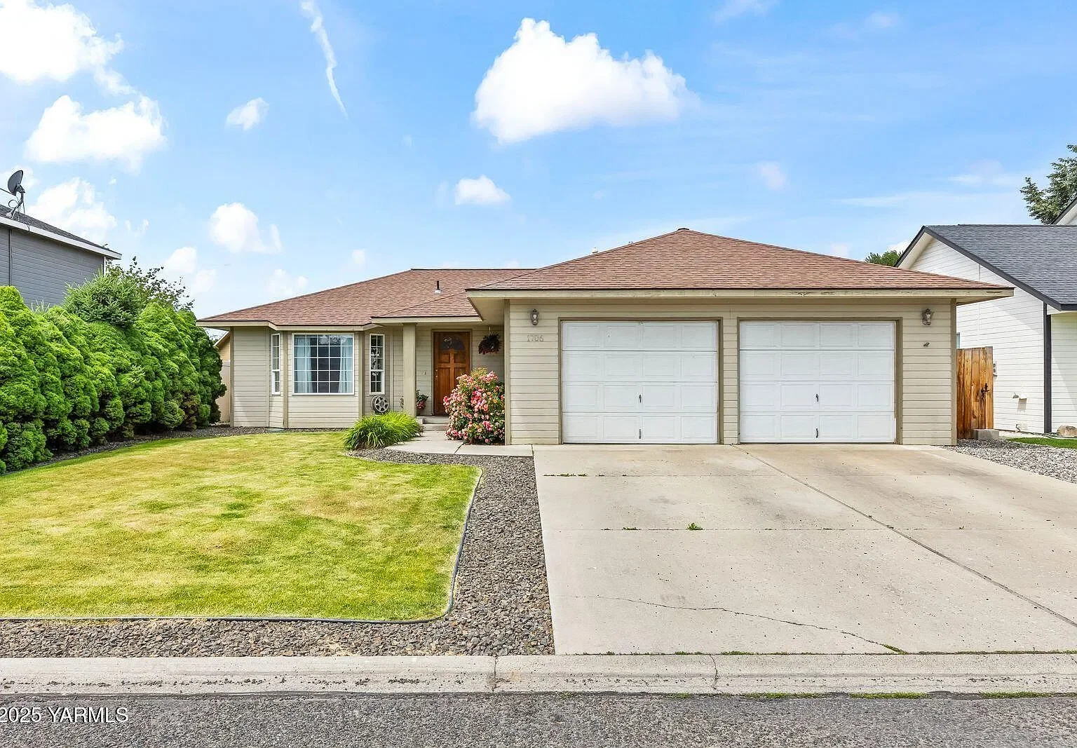 Single-story house with beige siding, a double garage, brown roof, front lawn, and pink flowering shrub near the front door, under a partly cloudy sky.