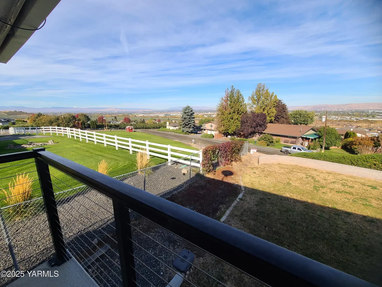 View of a suburban neighborhood from a balcony, showing a grassy yard with a white fence, trees with colorful foliage, a house with a brown roof, parked cars, and a clear sky with some clouds.