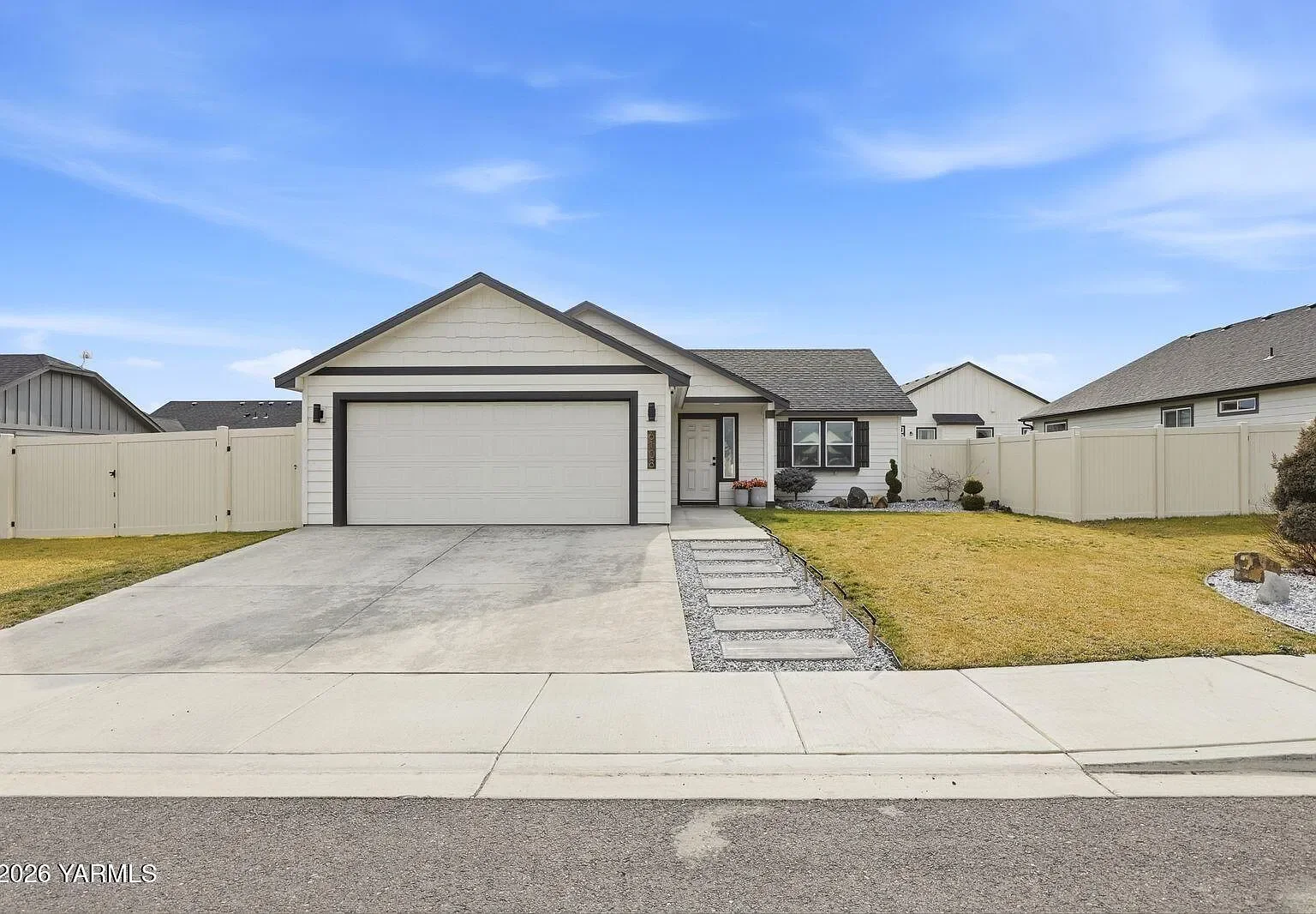 Front view of a modern single-story house with a white garage door, white exterior walls, and a small front yard with grass and decorative bushes, surrounded by a white fence.
