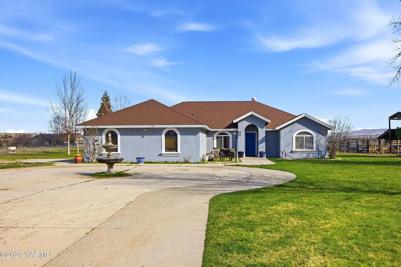 A single-story house with a blue exterior and a red roof, featuring a front door painted blue, arched and rectangular windows, surrounded by a yard with green grass, trees, potted plants, and a circular concrete driveway, with a blue sky above.