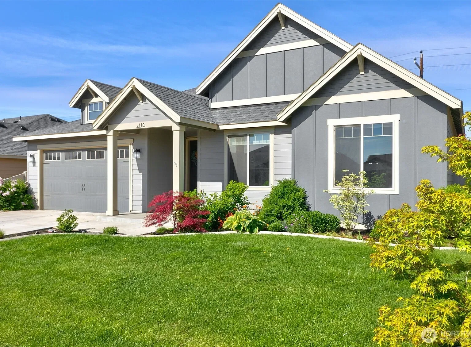 A modern two-story house with a gray exterior, large front windows, a attached garage, and a well-maintained front lawn with colorful bushes and a small tree.