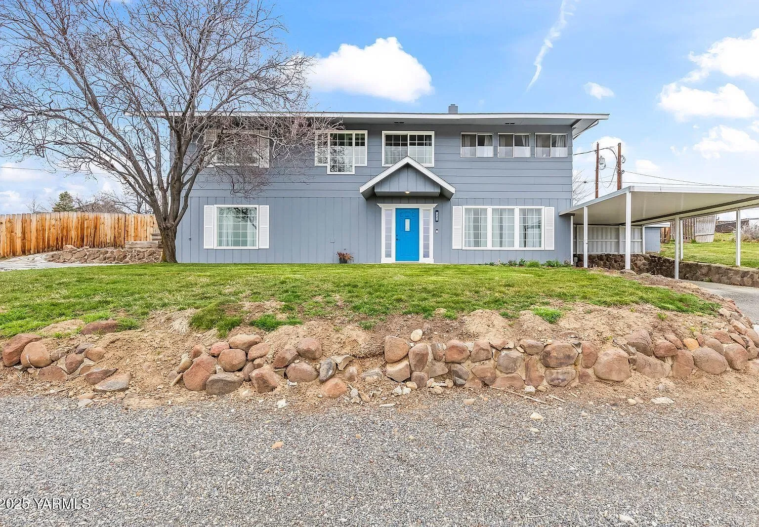 A two-story house painted in light blue with white window trims and a bright blue front door. The house has a sloped roof and multiple windows on both levels. There is a large leafless tree in front, and a grassy yard with some rocks along the border. To the right, there is a covered driveway or carport built with white support columns. A wooden fence surrounds part of the yard on the left side.