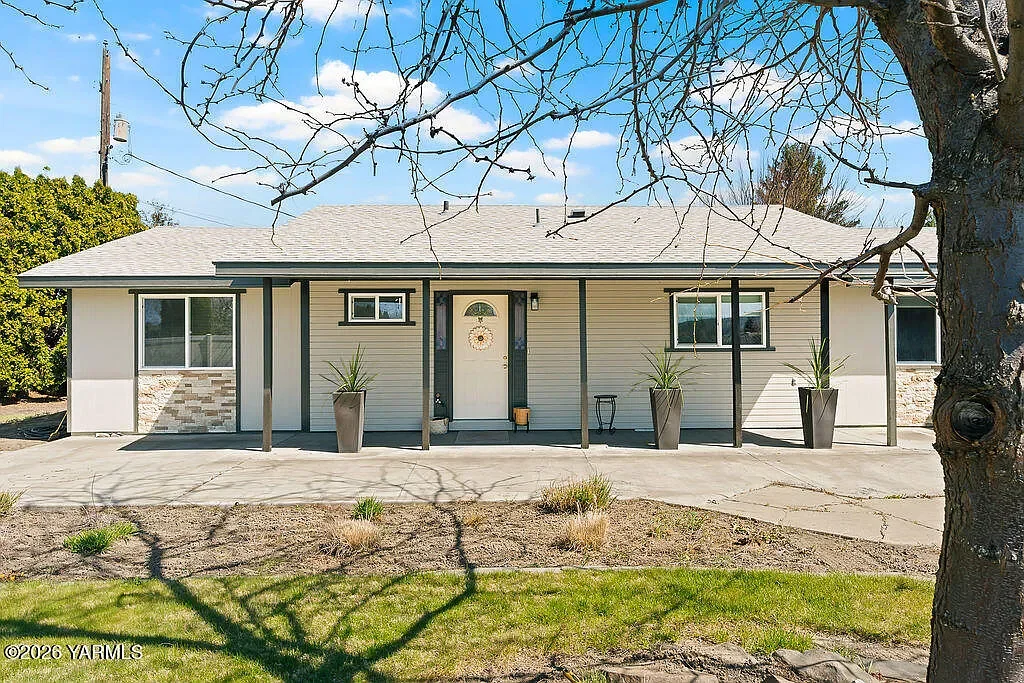 Front view of a single-story house with a light-colored exterior, gray shingles, and a small front porch. The porch has three large potted plants, a small table, and a door with a decorative wreath. Leafless tree branches are in the foreground, and the sky is partly cloudy and blue.