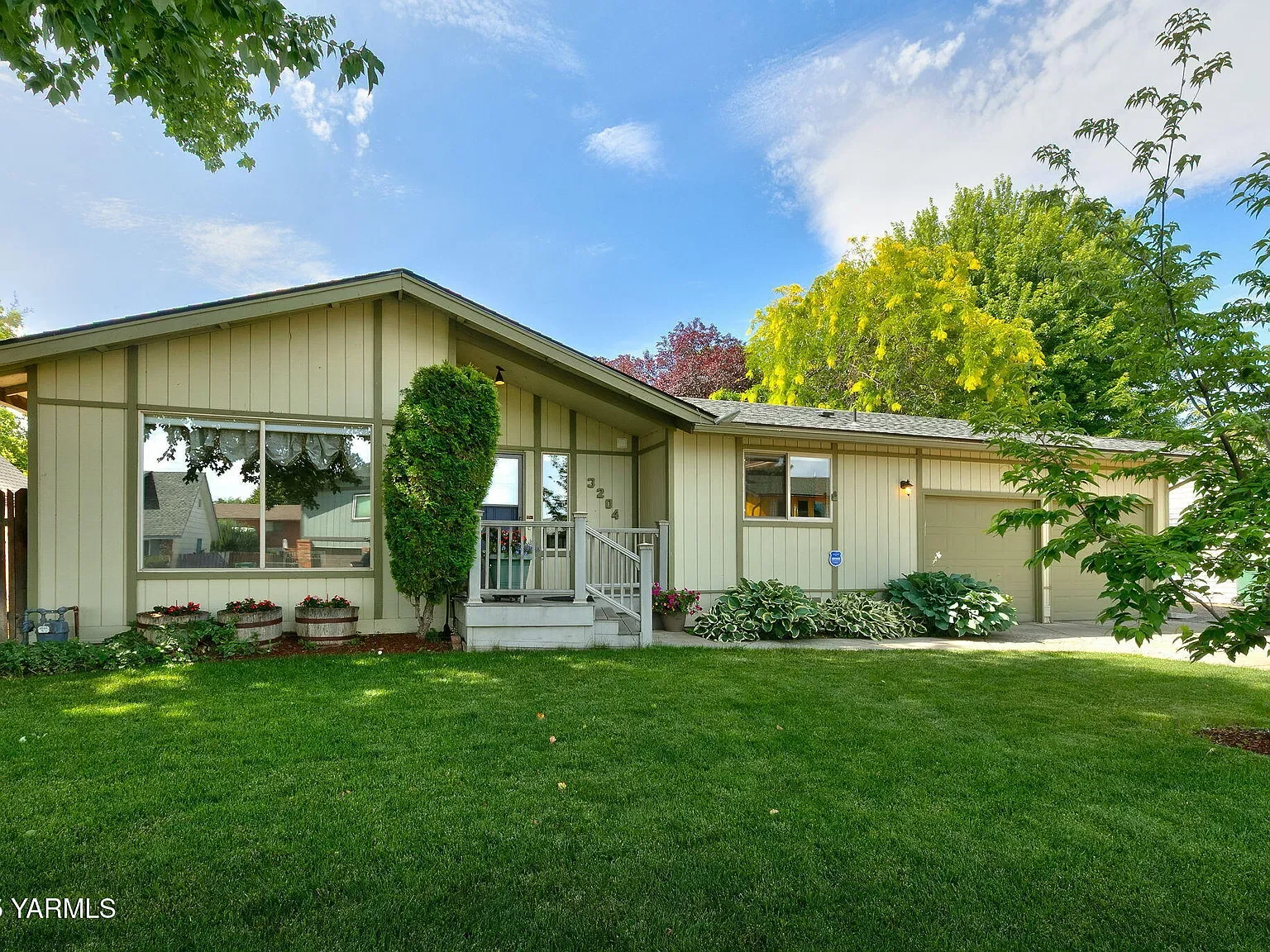 Light green single-story house with large front window, small porch, and attached garage. Front yard has lush green lawn, small bushes, and trees, with a partly cloudy blue sky overhead.