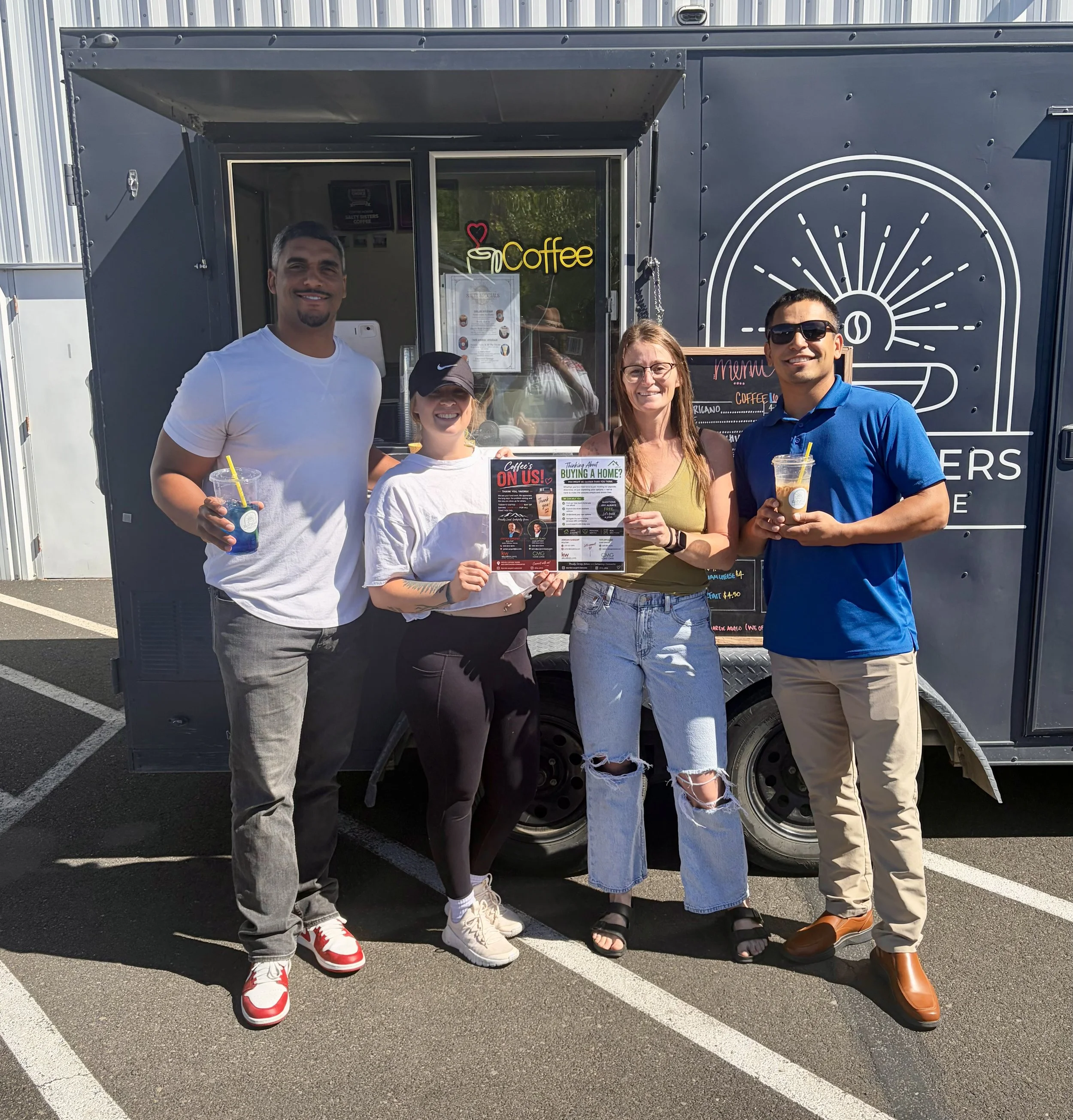 Five people standing in front of a black food truck, holding drinks, smiling, some wearing sunglasses, with a menu and sign visible on the truck.