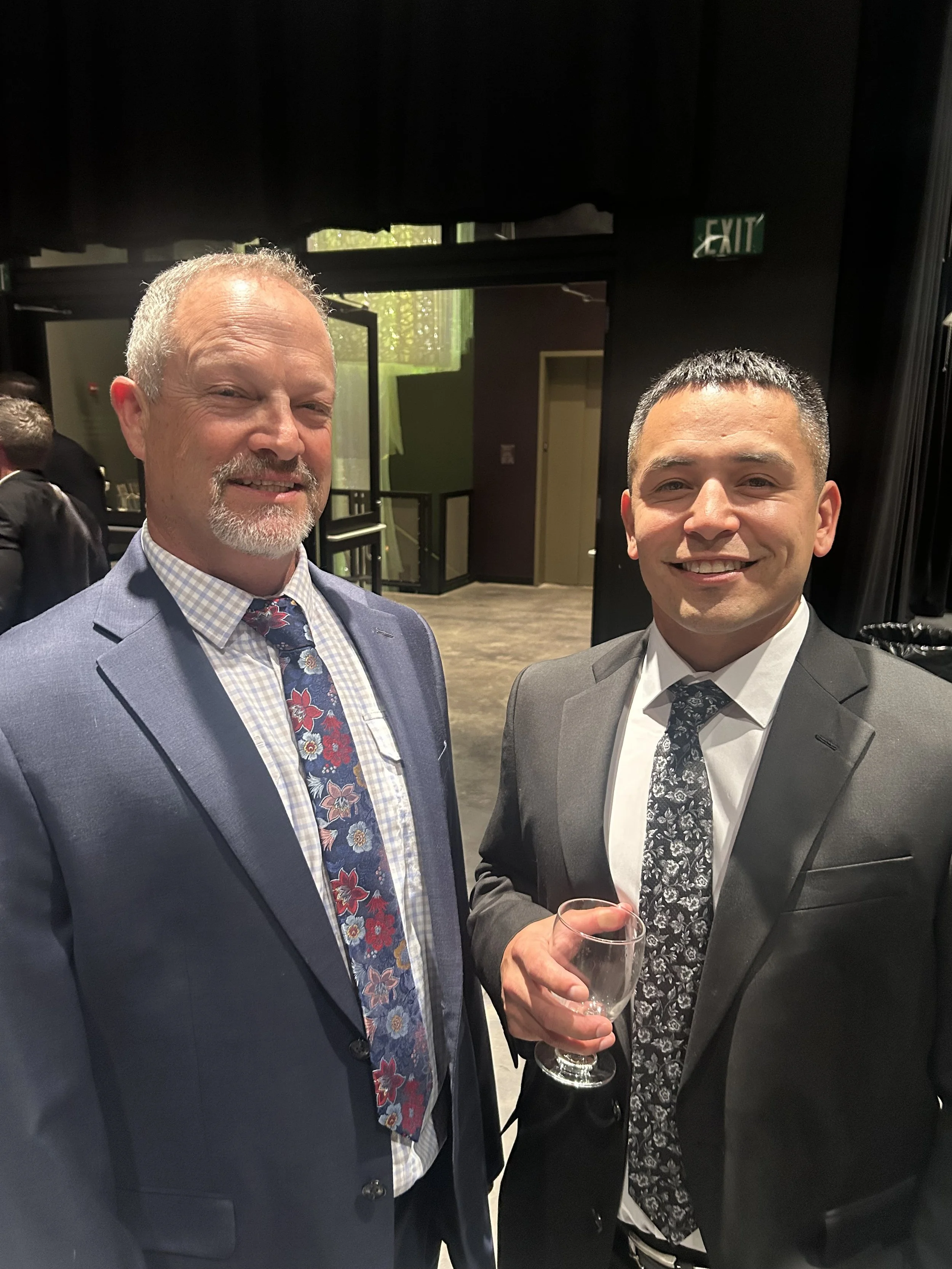 Two men dressed in suits at a social event, smiling at the camera. The man on the left has gray hair and a beard, wearing a blue suit and a floral tie. The man on the right has short dark hair, wearing a gray suit and a patterned tie, holding a wine glass. The background shows other attendees and an emergency exit sign.