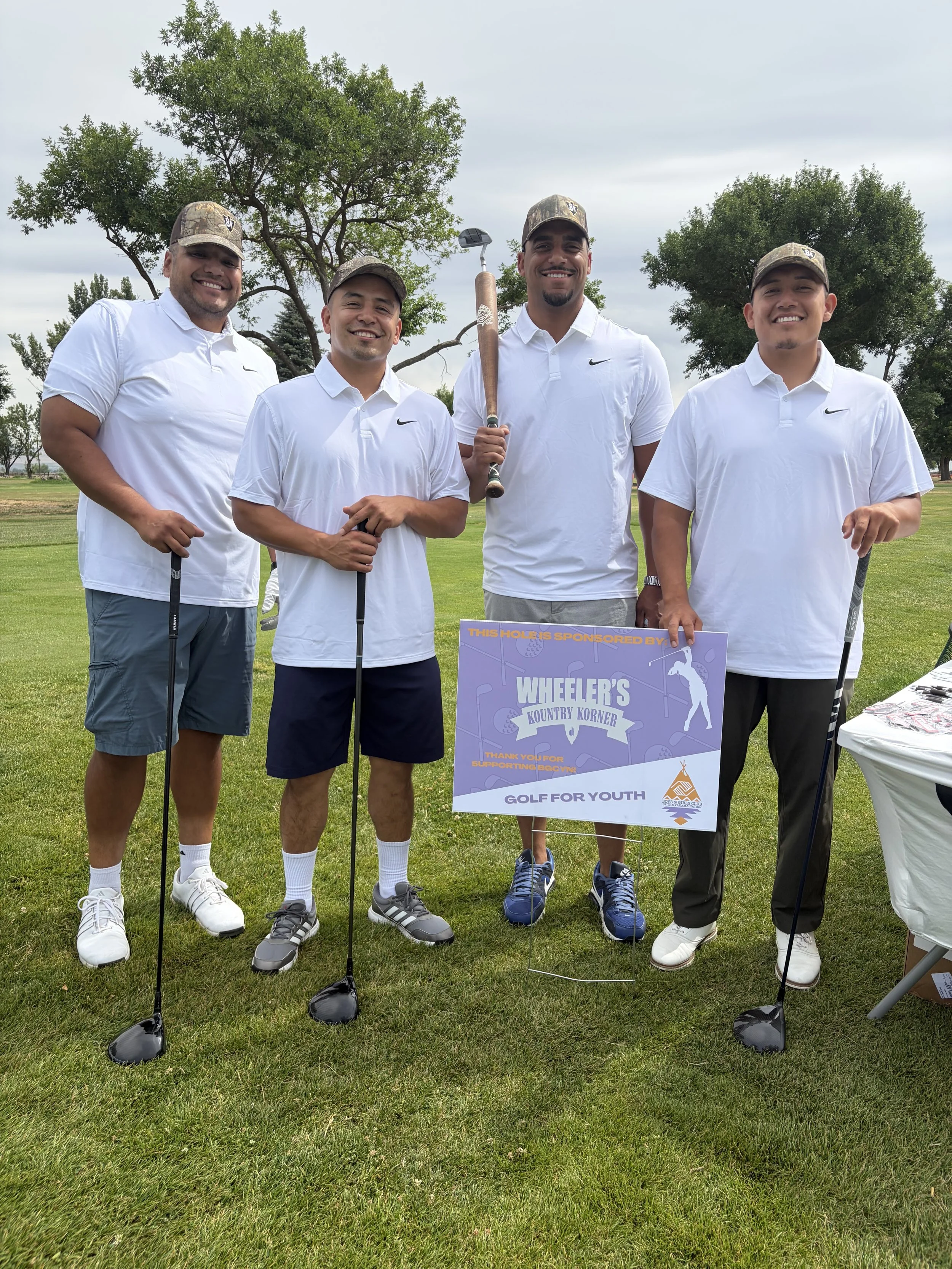 Four men standing on a golf course holding golf clubs, smiling, wearing white shirts and camouflage caps, with trees and cloudy sky in the background.