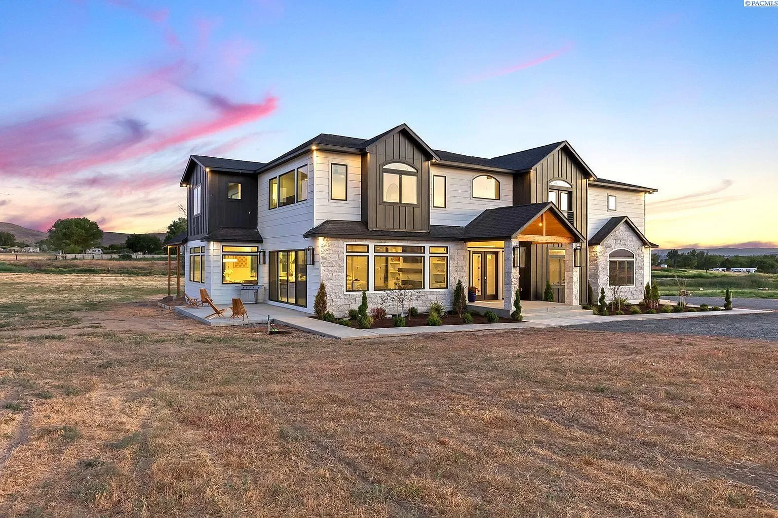 Modern two-story house with white and dark gray exterior, large windows, landscaped front yard, and dirt lawn at sunset.