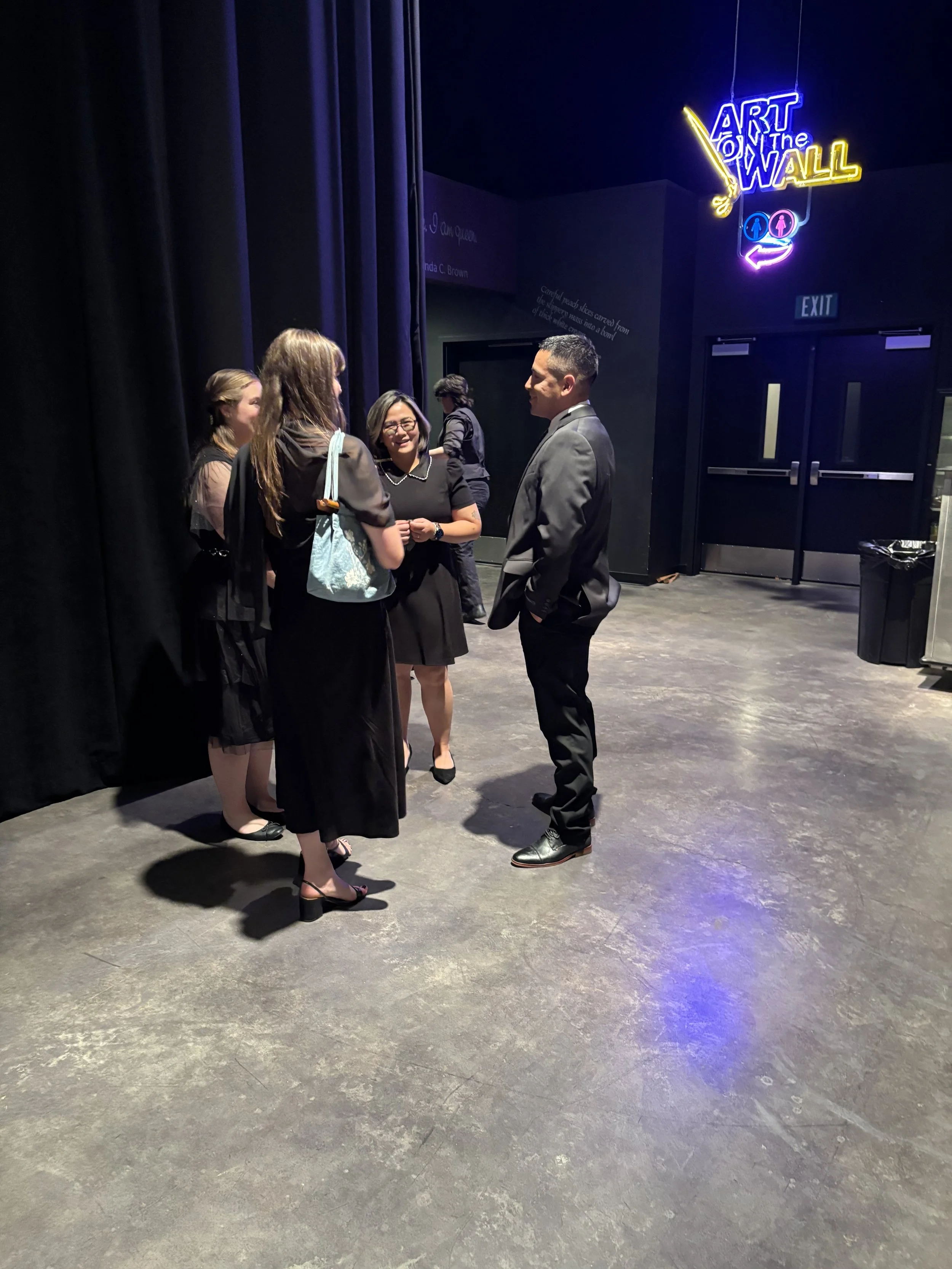 Four women and one man dressed in formal attire standing in a circle talking inside a dark room with purple and yellow neon signage that reads 'Art on the Wall' and restroom symbols.