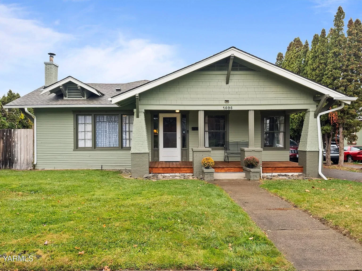 Front view of a light green house with a wooden porch, three steps, and flower pots, surrounded by green grass and trees.
