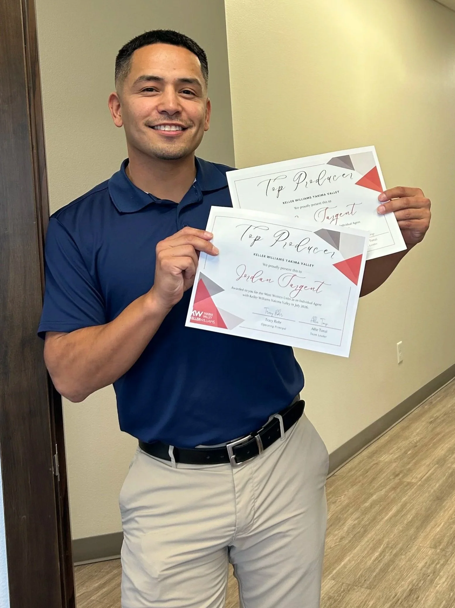 A man in a blue shirt and beige pants holding two certificates in a hallway.