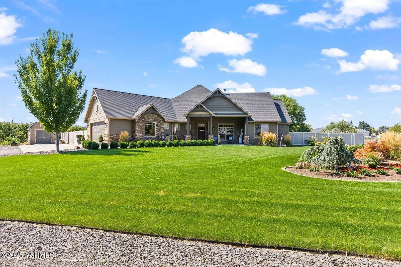 A large, modern house with a well-maintained green lawn in front. The house has a mix of stone and siding exterior with multiple gables and a front porch. There are landscaped bushes, trees, and flower beds around the yard, and a bright, partly cloudy sky overhead.
