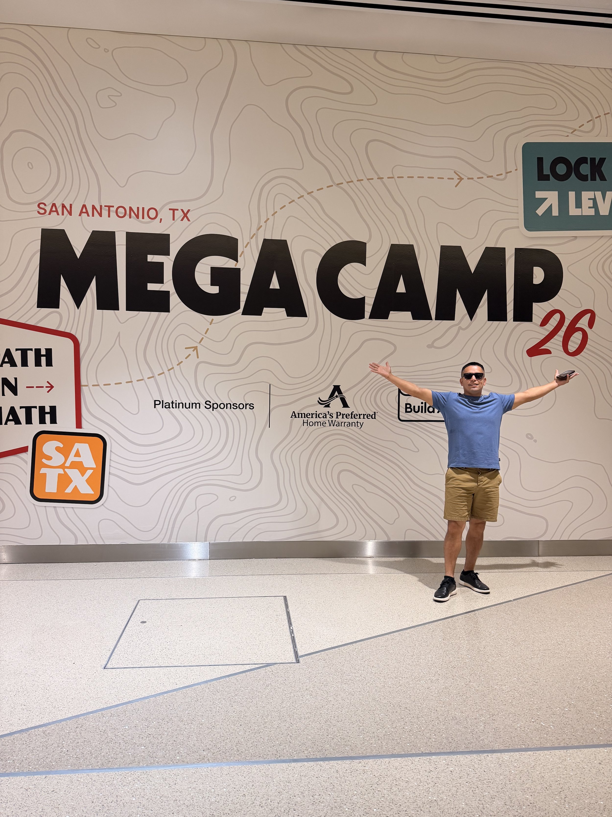 A man standing with arms outstretched in front of a large map poster at an airport, with the words 'Mega Camp' and 'San Antonio, TX' visible on the poster.