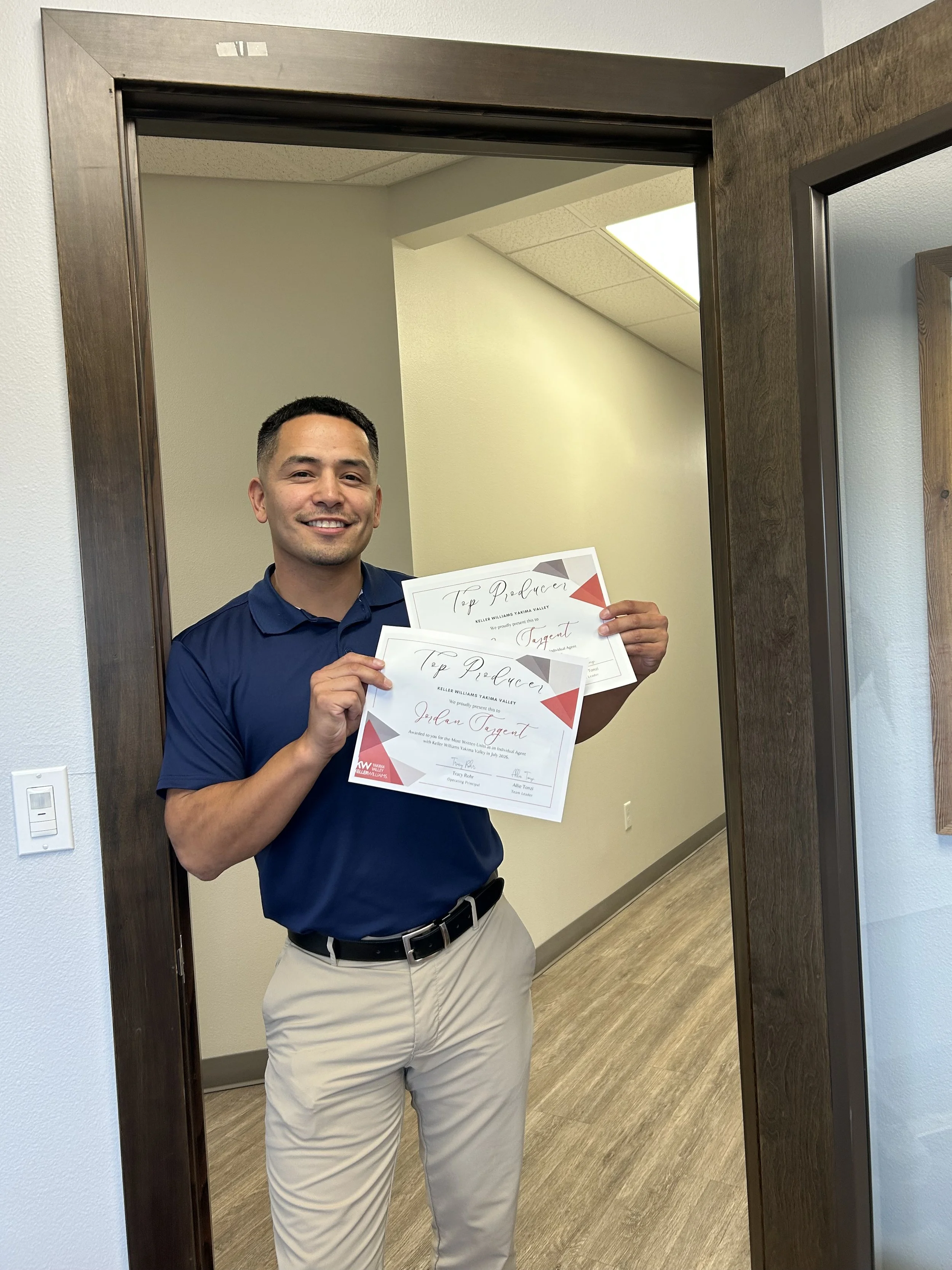 A man smiling and holding two certificates of achievement in front of a doorway. He is wearing a navy blue polo shirt and beige pants.