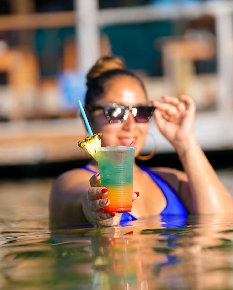 A woman in sunglasses holding a colorful layered drink with a pineapple slice garnish while partially submerged in water, smiling at the camera.