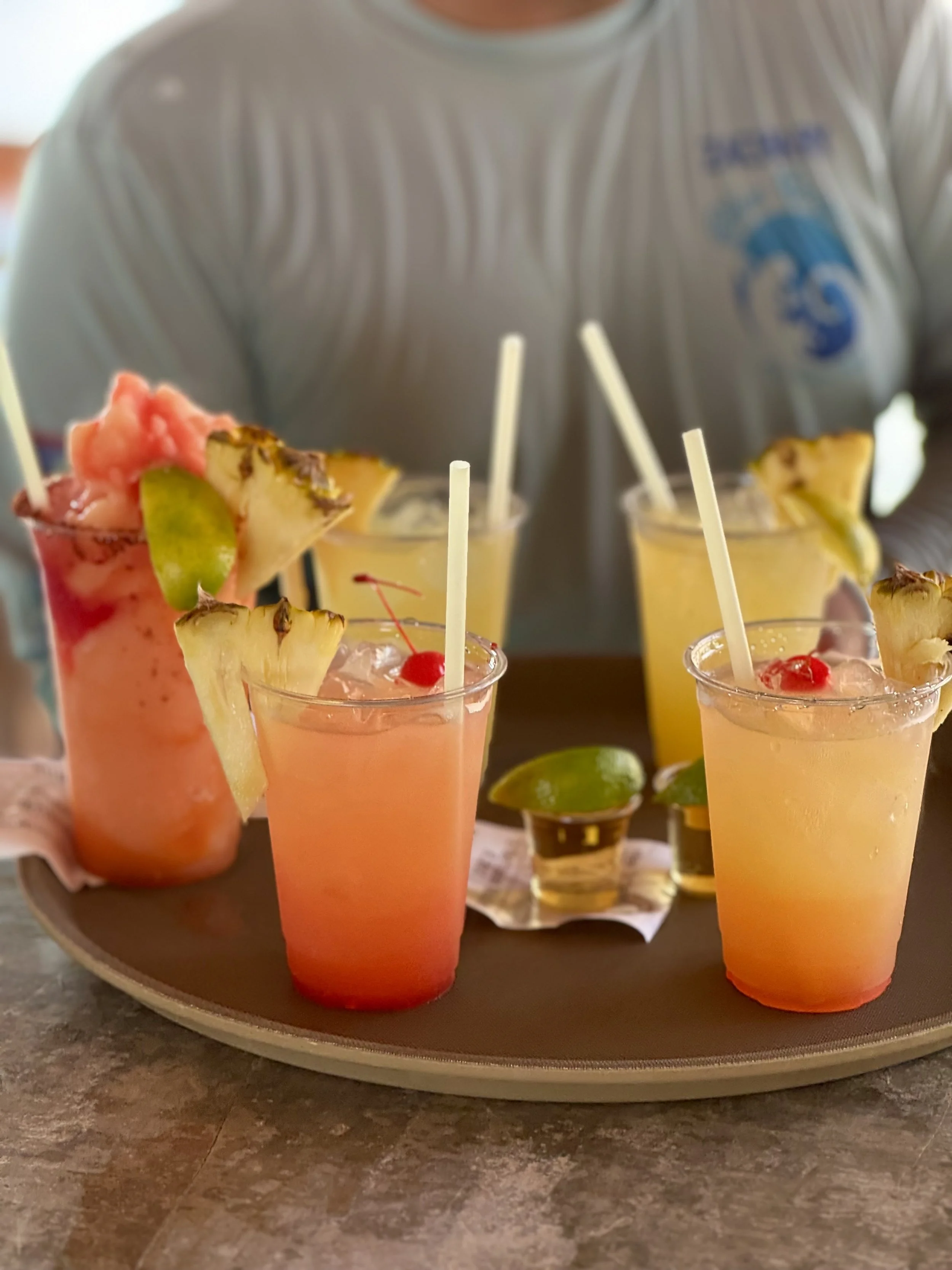 Assorted tropical fruit cocktails with garnishes on a round silver tray, on a wooden surface.