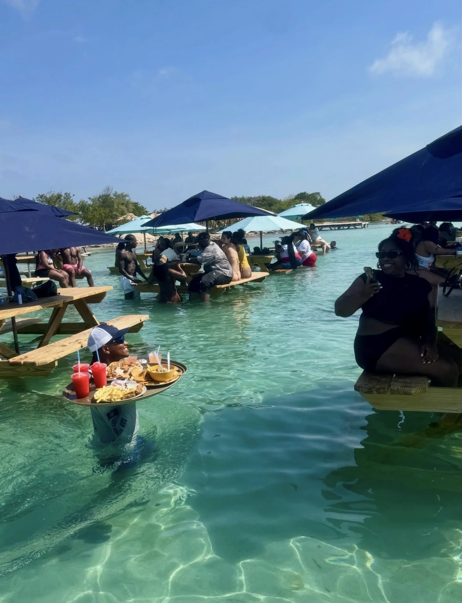 People enjoying a ocean with umbrellas, some sitting on tables in the water, and a person holding a tray of food and drinks.