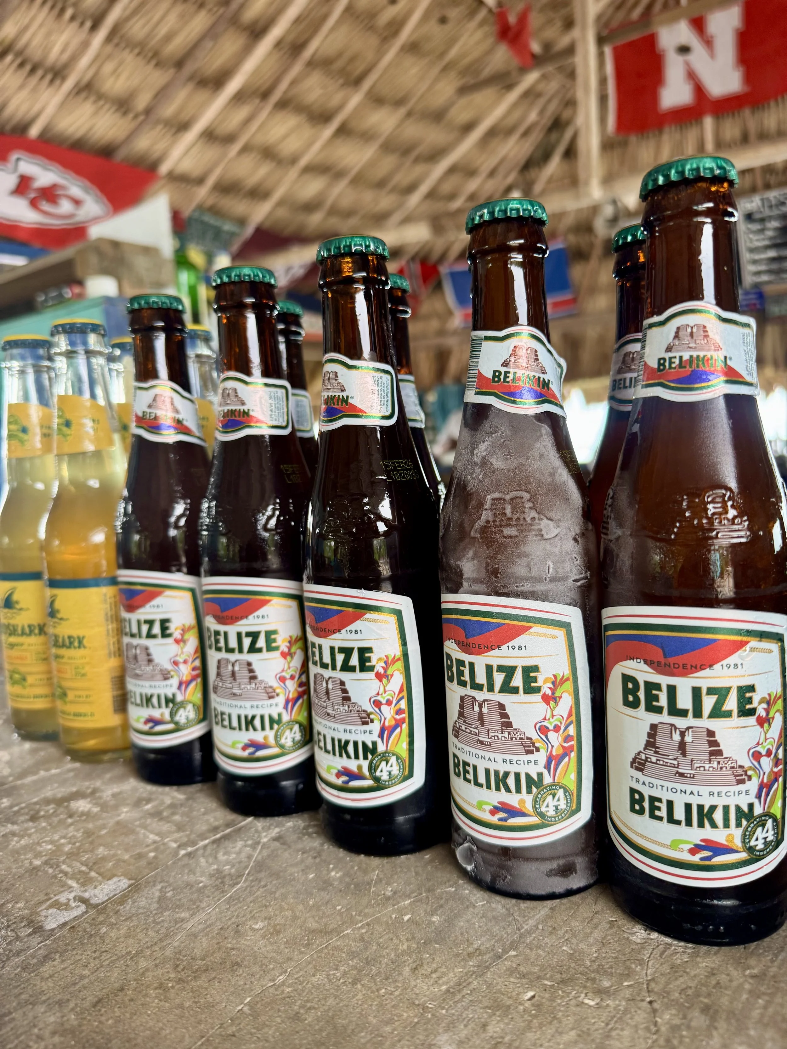 A row of bottled beverages, including Belikin beer, lined up on a wooden surface with a thatched roof and colorful banners hanging in the background.