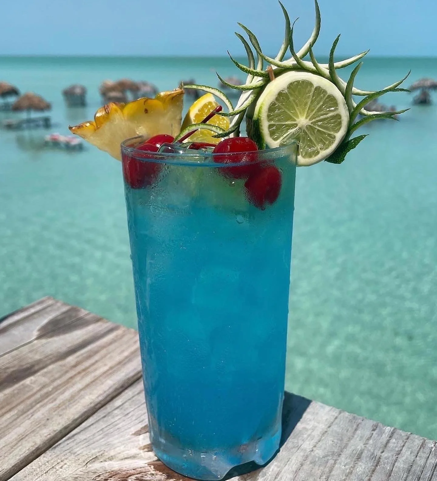A tall glass of blue tropical cocktail garnished with lemon, pineapple, and cherry, placed on a wooden surface near a beach with turquoise water and umbrellas in the background.