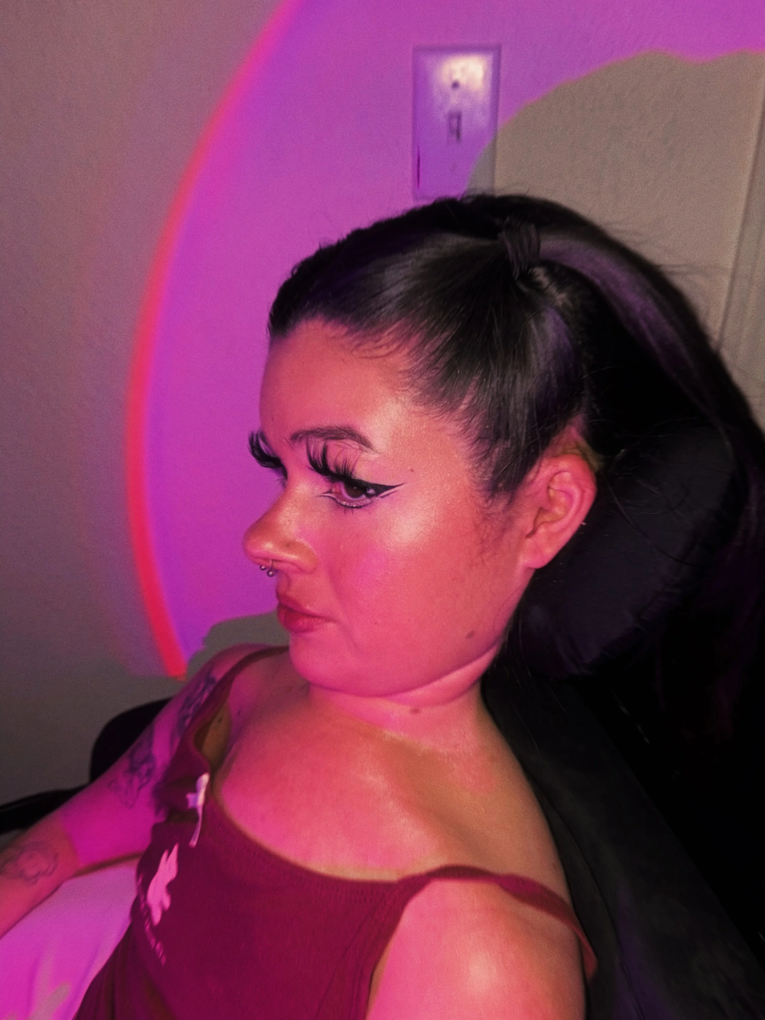 Close-up profile of a woman with dark hair styled in a braided ponytail, with makeup including winged eyeliner, in a room illuminated with pink and purple lighting. She is wearing a purple lace top and has tattoos on her arm.