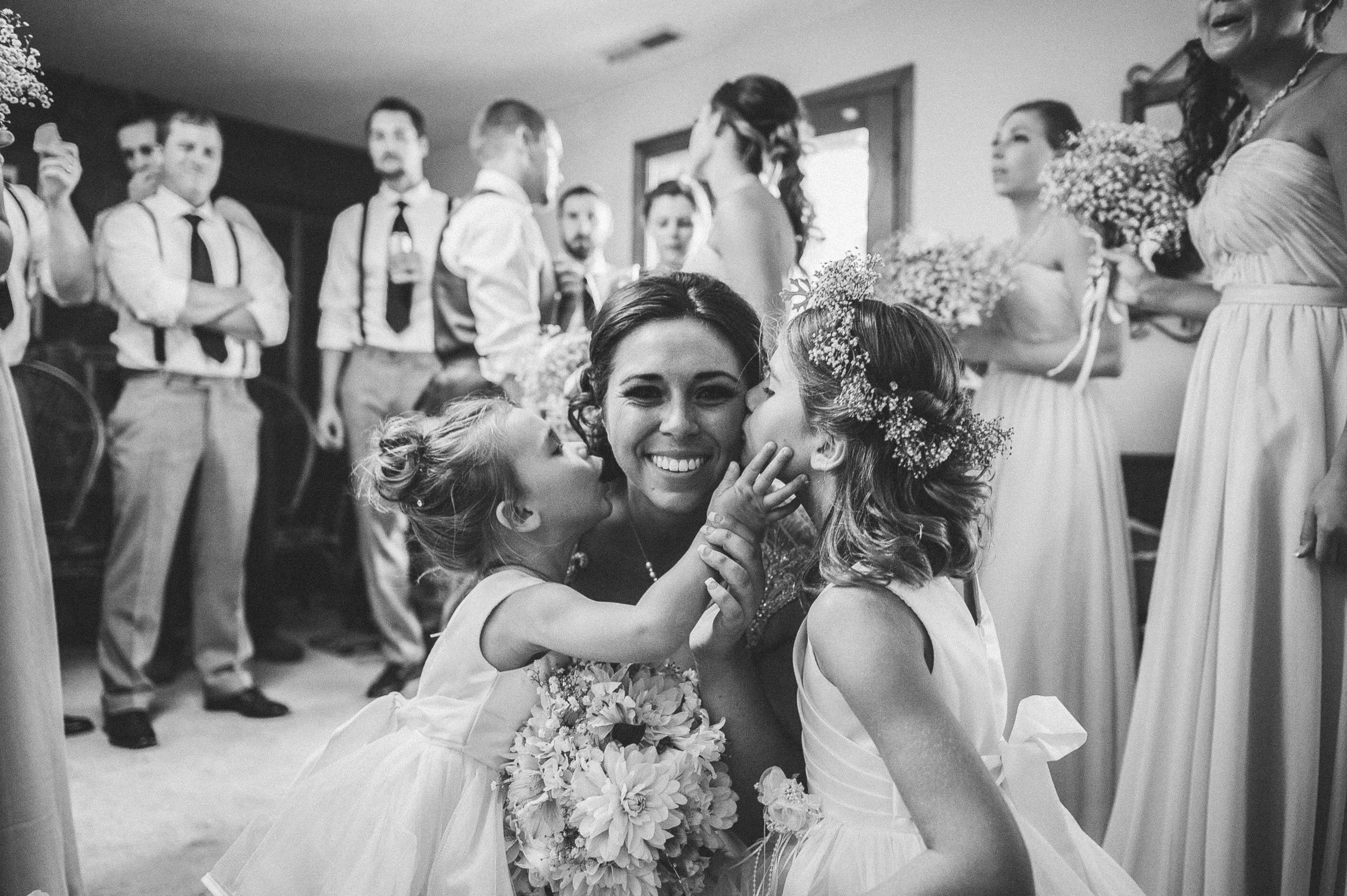 Black and white photo of a wedding scene with a bride and young girl facing each other, smiling and touching faces. Other women in dresses holding flower bouquets and men in shirts with suspenders are in the background.