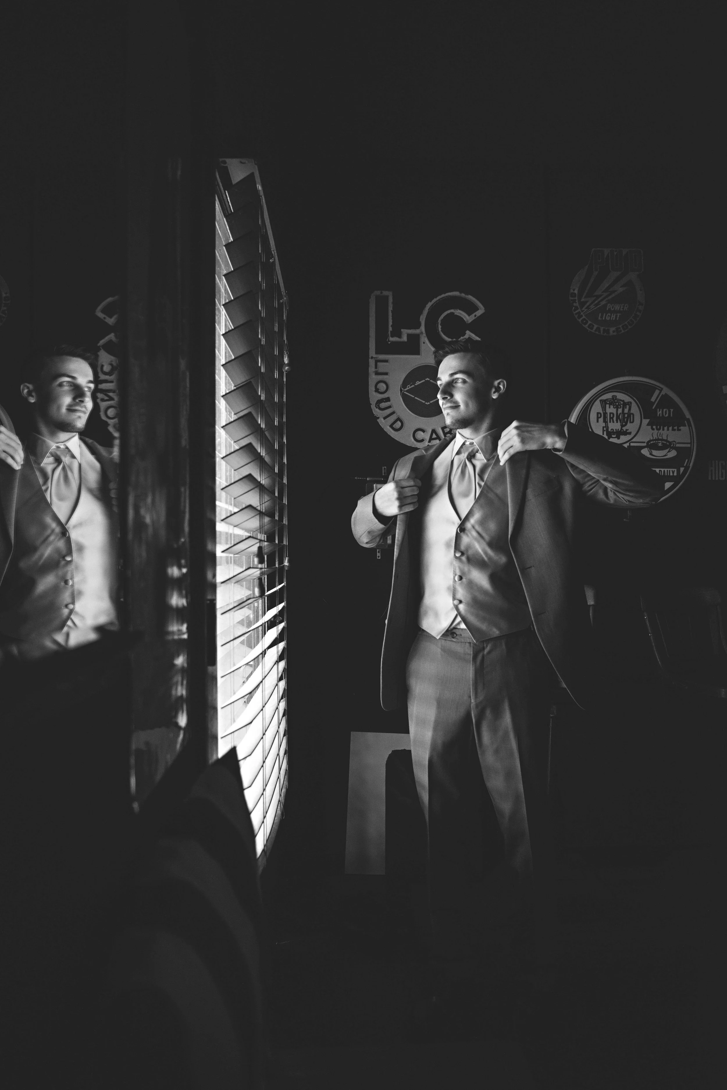 A man in a suit standing in front of a mirror, adjusting his jacket, with various signs on the dark wall behind him, including the logos for liquor and food companies.