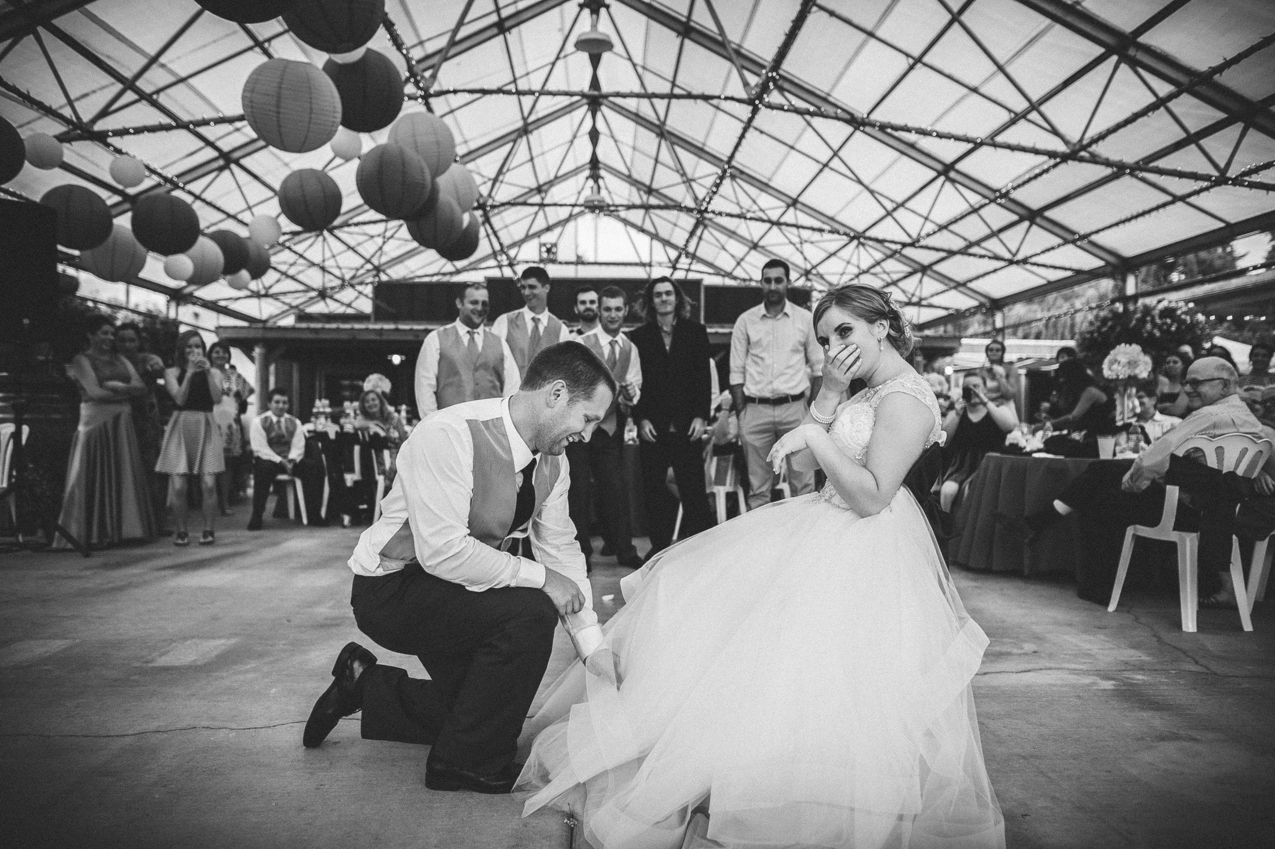 A black and white photo of a wedding reception, showing a bride and groom dancing. The groom is kneeling and holding the bride's hand, who is wearing a wedding gown and appears to be laughing or surprised. Guests in formal attire are gathered around watching, with some seated at tables and others standing. The venue has a glass or transparent roof with decorative paper lanterns hanging.