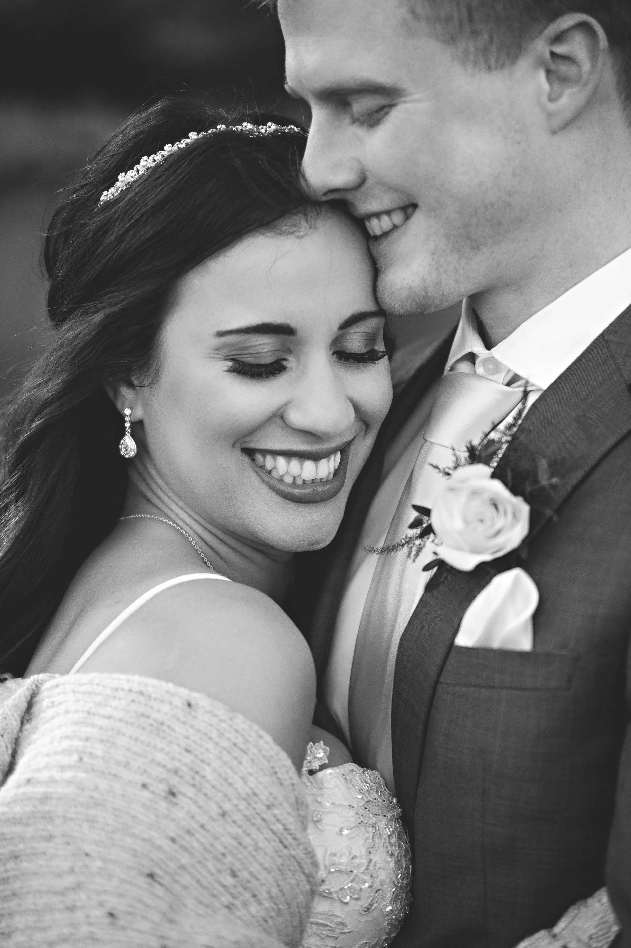 Black and white photo of a bride and groom smiling and embracing during their wedding. The bride has dark hair, a beaded headband, and earrings; the groom is wearing a suit with a boutonniere.