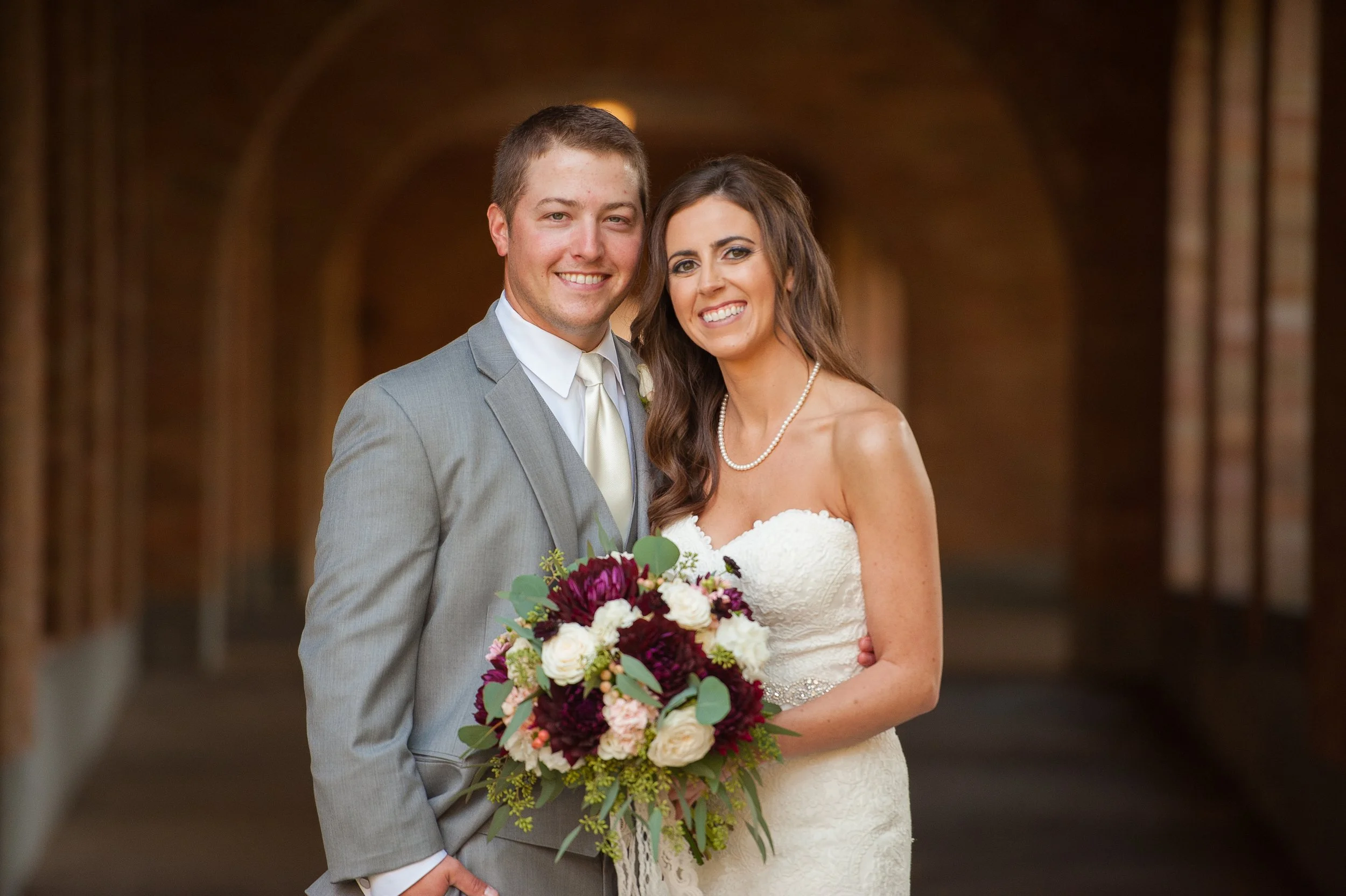 A happy couple at their wedding, with the groom in a gray suit and the bride in a white strapless dress, holding a bouquet of purple, white, and pink flowers, standing in a covered outdoor walkway.