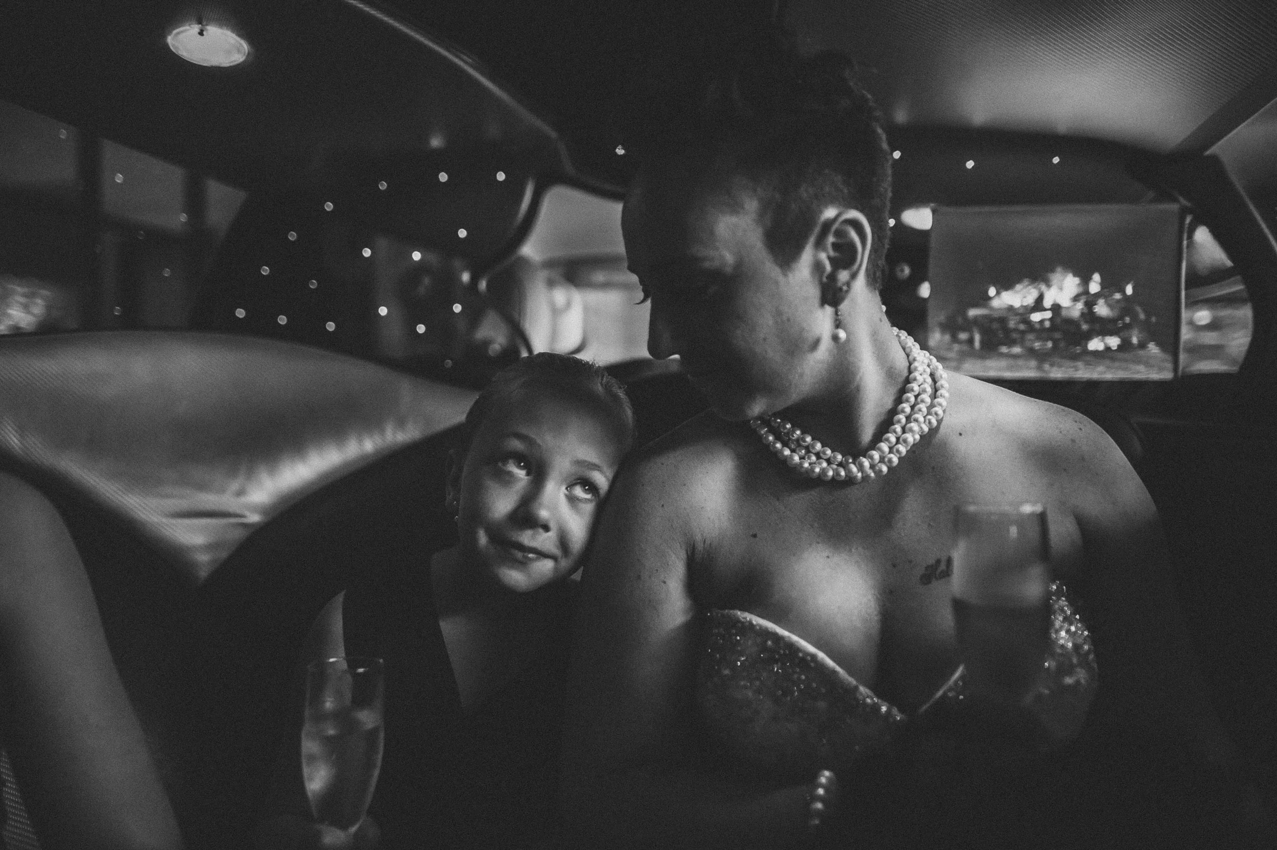 A woman and a young girl sit close together in the back of a vehicle, looking at each other. The woman is wearing multiple strings of pearls and has short hair, while the girl has curly hair and is gazing upward at the woman. The vehicle's interior and window reflections are visible in the background.