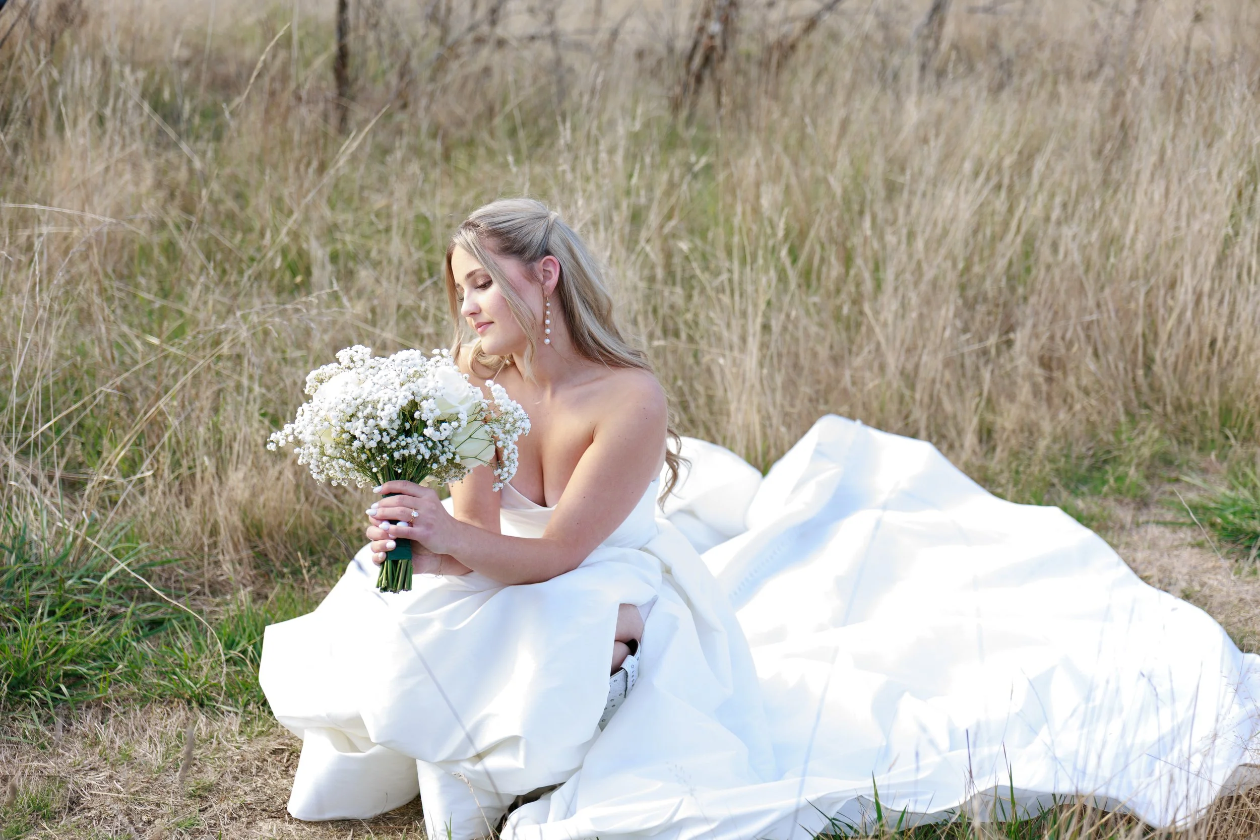 A woman in a white dress sits on grass in a field, holding a bouquet of white flowers and looking at them with a gentle expression.